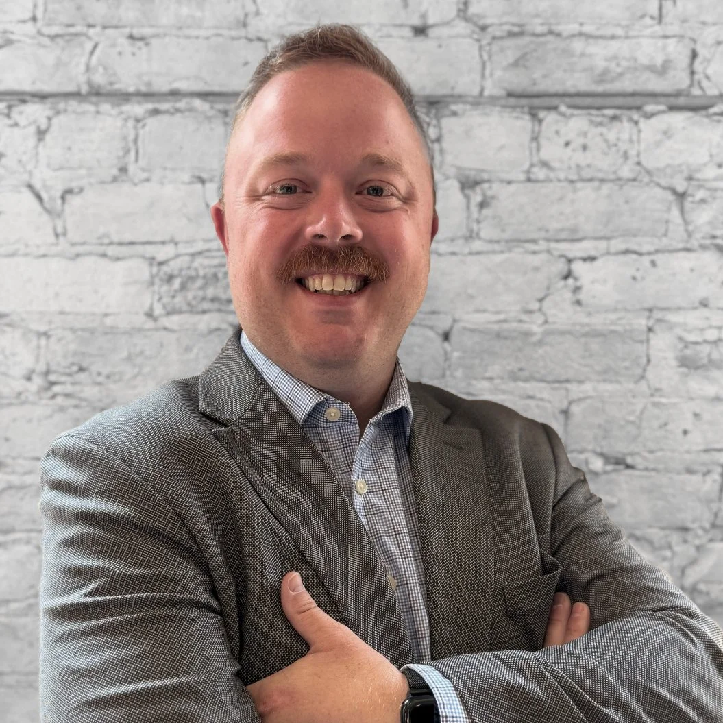 A smiling man with a mustache and short hair, wearing a gray suit and a checkered shirt, standing in front of a white brick wall.