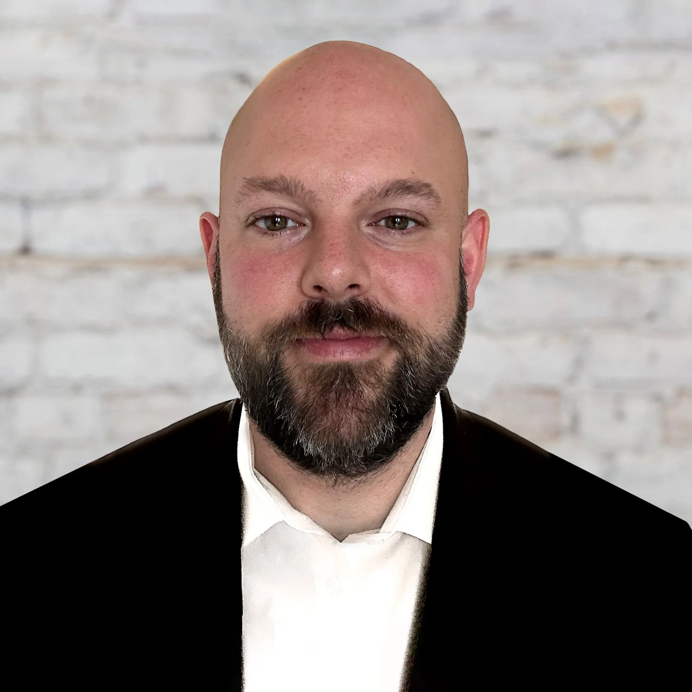 Portrait of a bald man with a beard, wearing a black blazer and white shirt, standing in front of a white brick wall.
