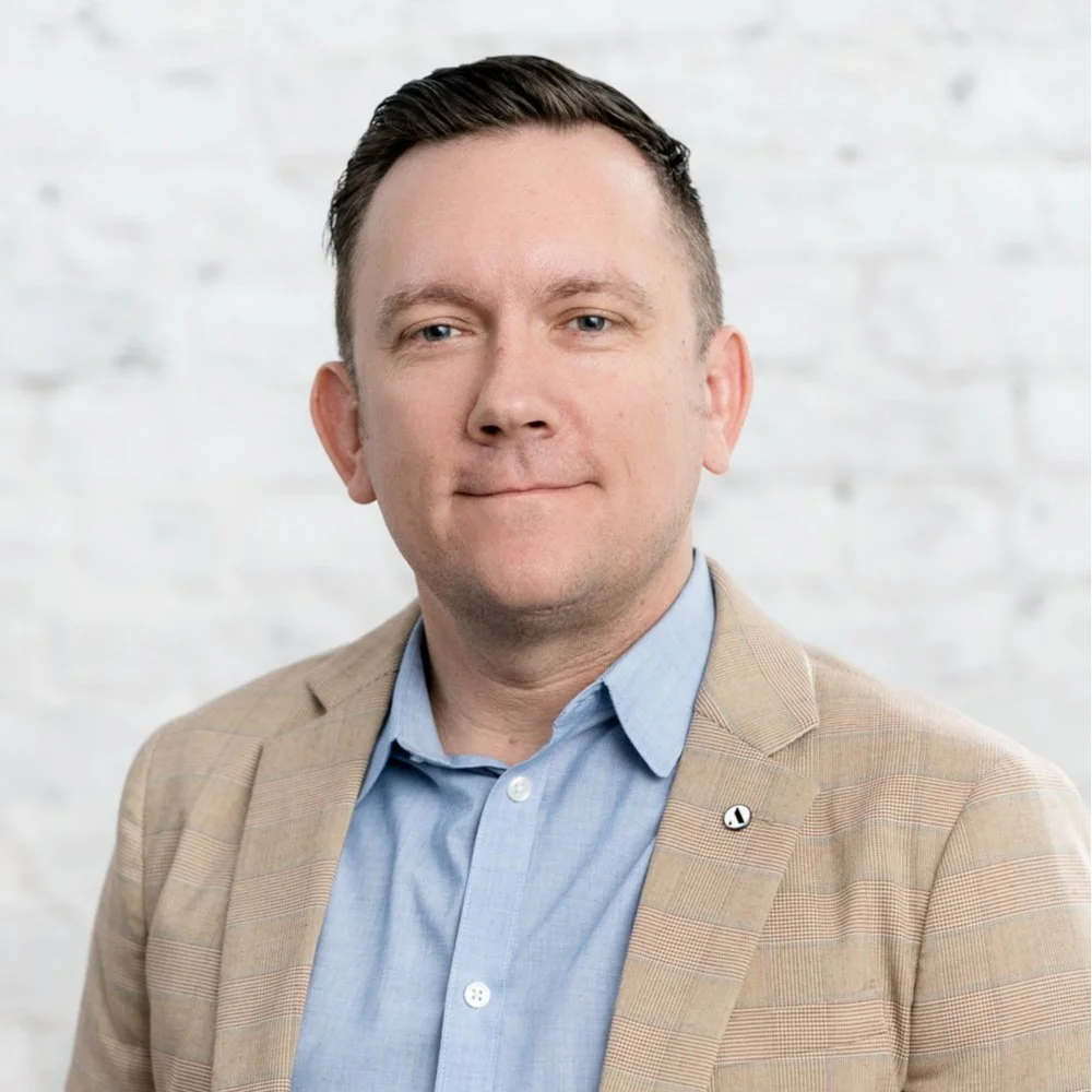 Headshot of a man with short dark hair, wearing a tan blazer and light blue dress shirt, standing against a white brick wall background.
