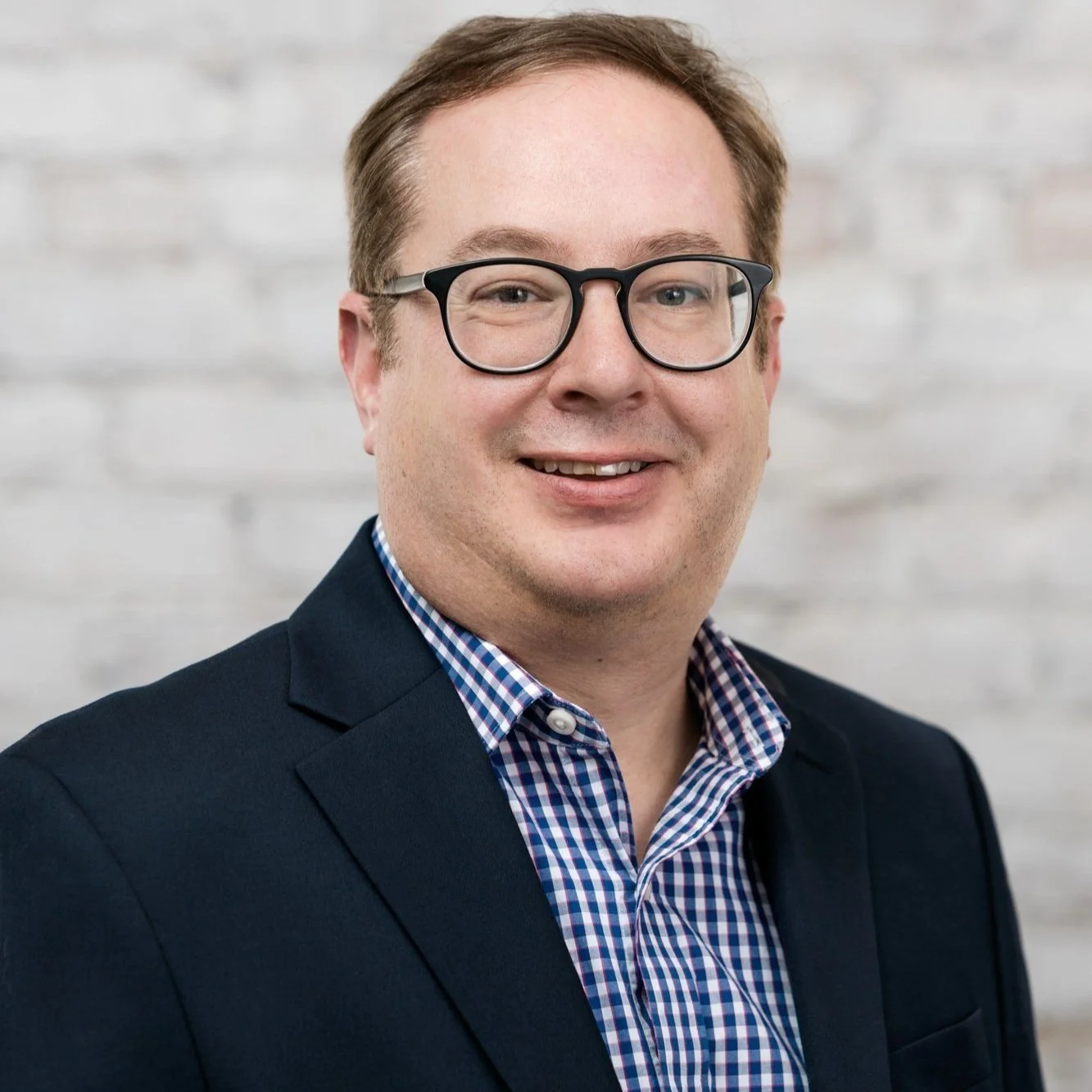 Portrait of a smiling man with glasses, wearing a dark blazer and a checkered shirt, posed against a light-colored brick wall.