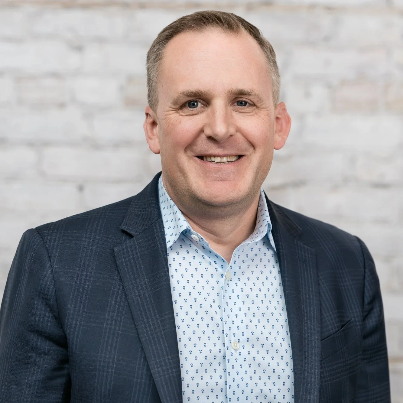 Portrait of a smiling man in business attire standing in front of a white brick wall.