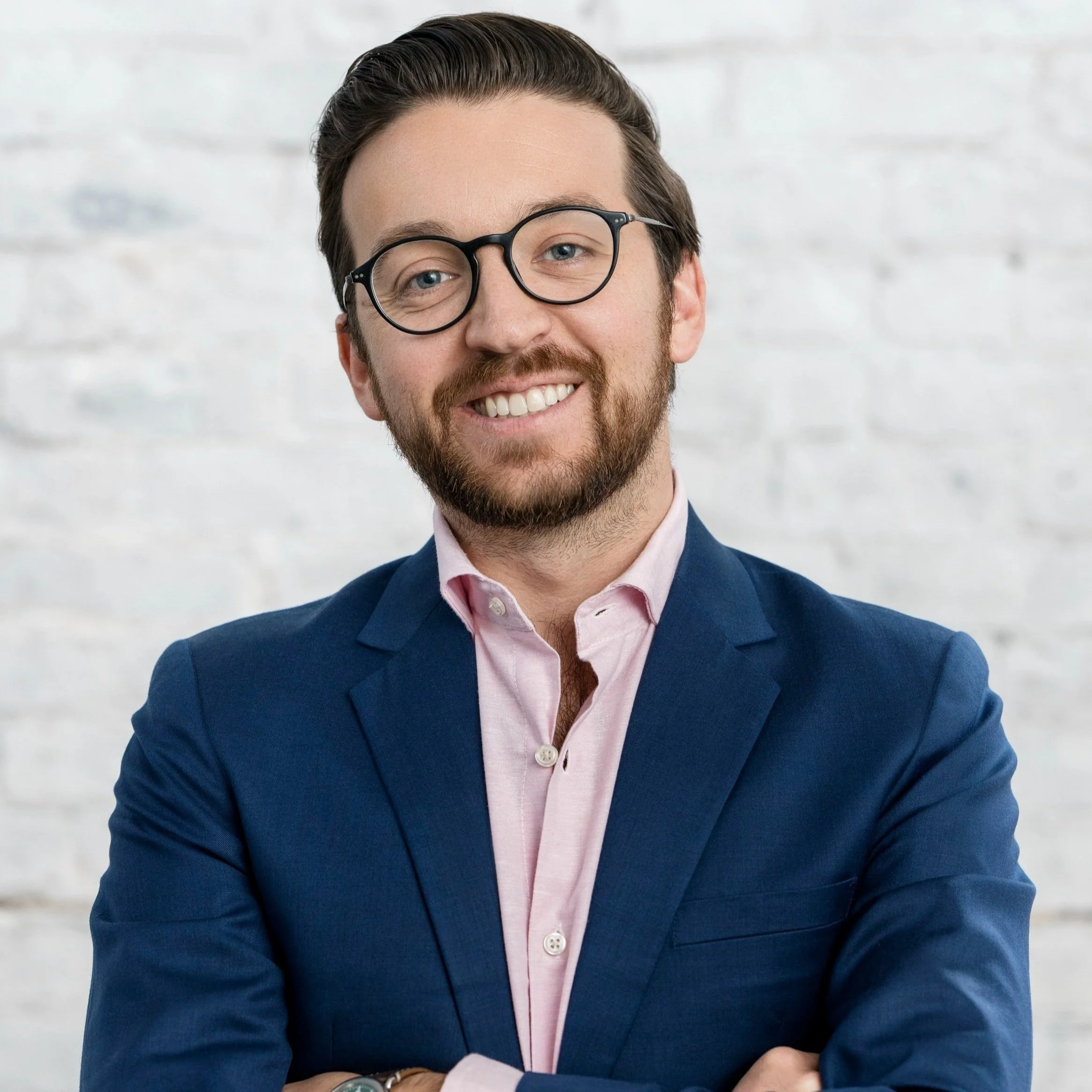 A man wearing glasses and a blue blazer smiling with crossed arms against a white brick wall.