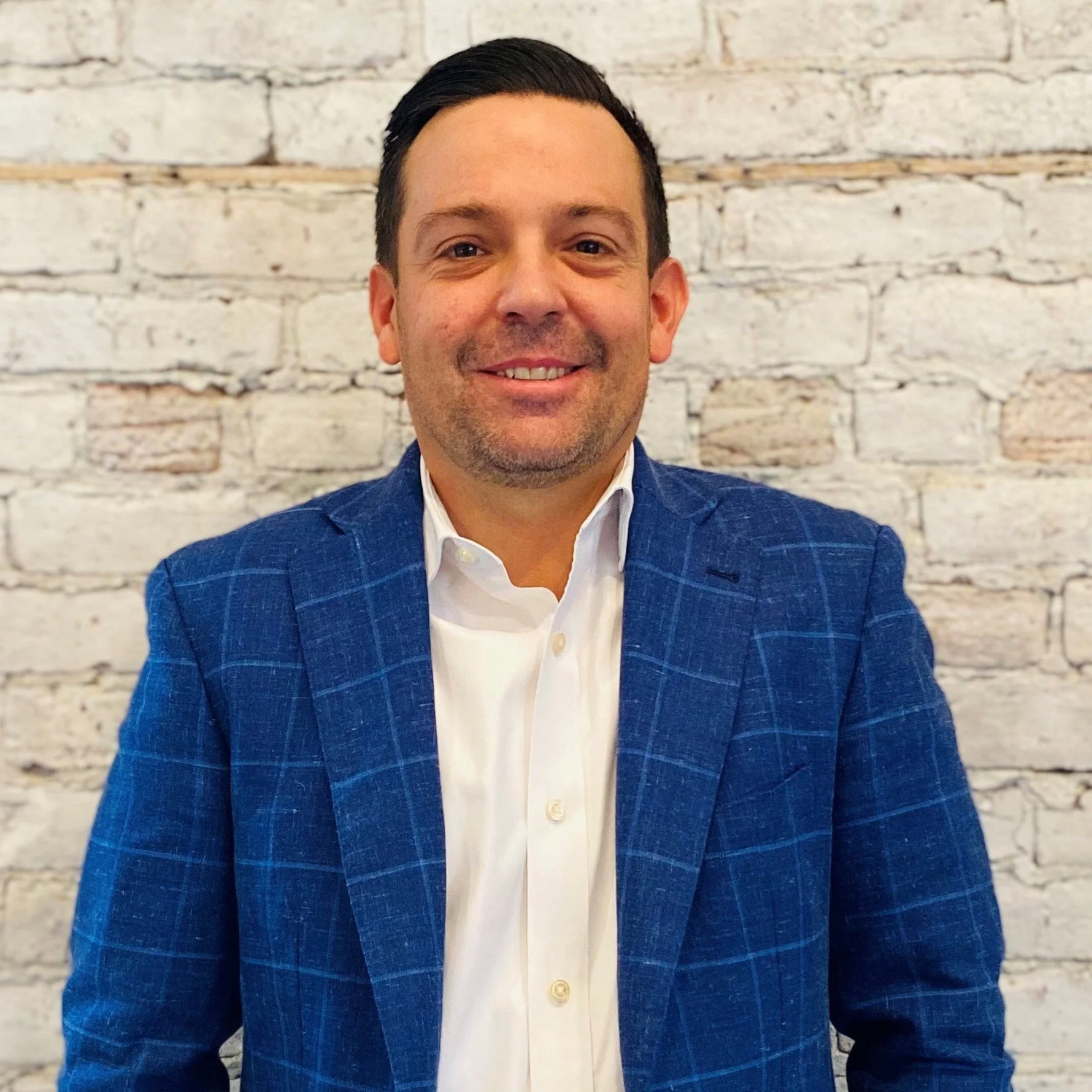 A man wearing a white shirt and blue blazer, smiling, with a white brick wall in the background.