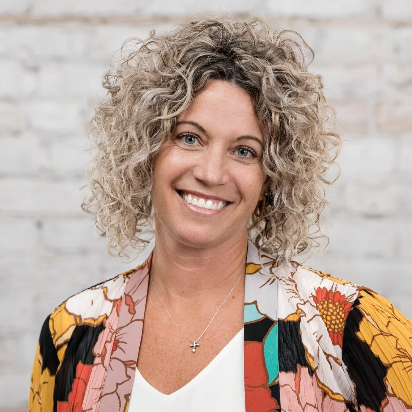 A woman with curly blonde hair smiling, wearing a colorful patterned blazer and a necklace with a cross pendant, standing in front of a light-colored brick wall.