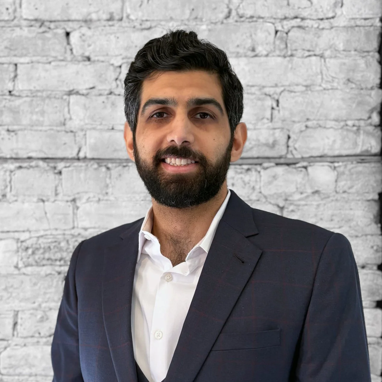 A man with dark hair and a beard wearing a dark suit jacket and white shirt, standing in front of a gray brick wall.