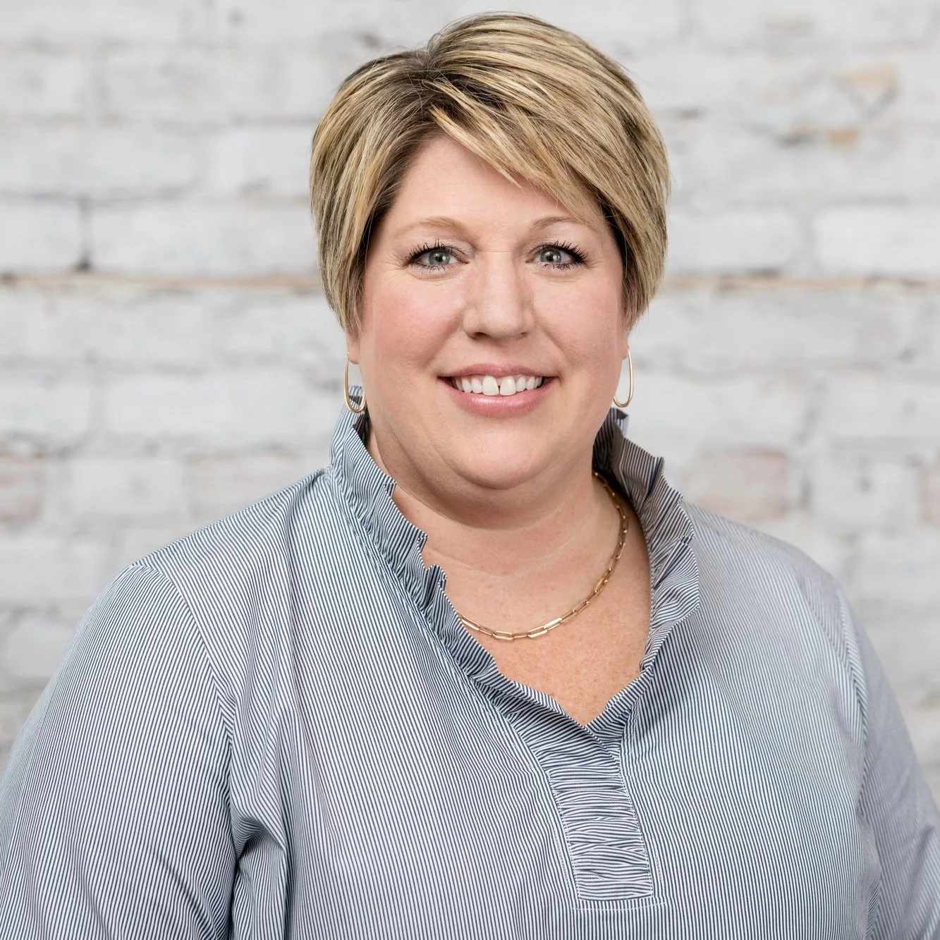 A woman with short blonde hair smiling, wearing a light blue striped blouse and gold jewelry, standing in front of a white brick wall.