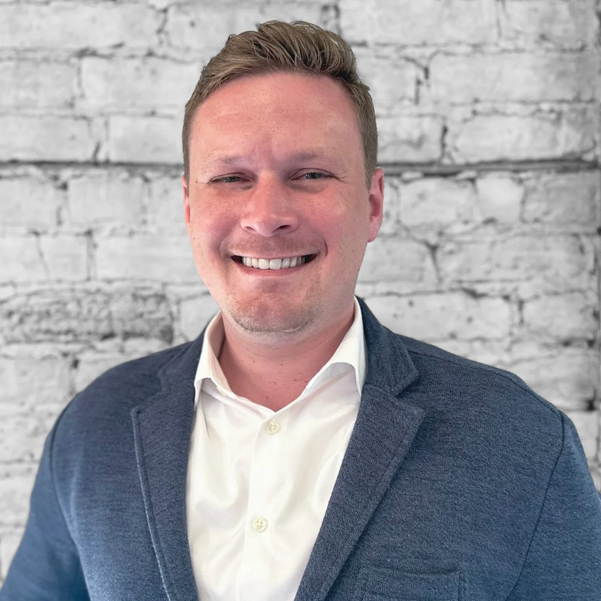 Portrait of a smiling man wearing a white shirt and a dark blazer, set against a white brick wall background.