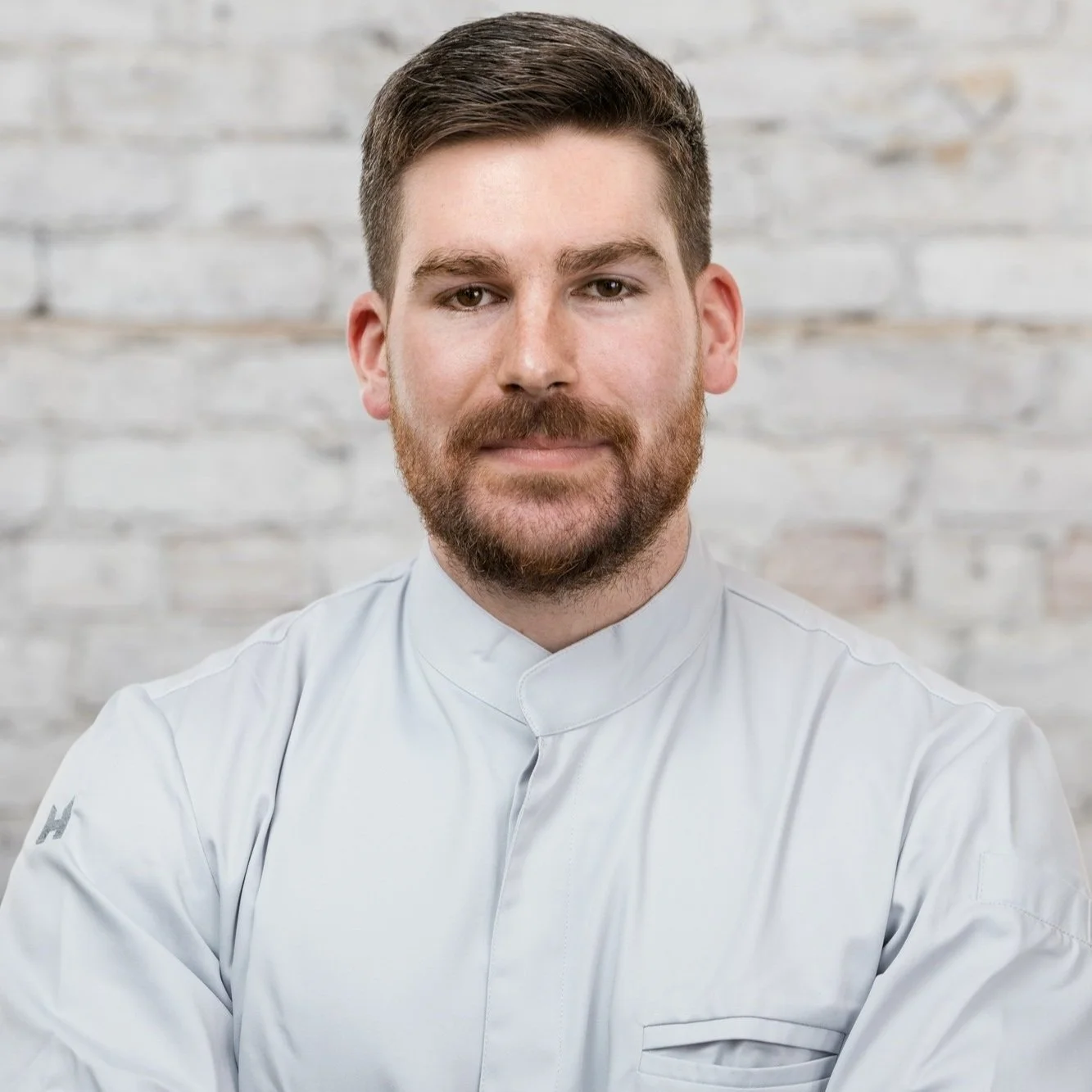 Portrait of a man with brown hair and beard wearing a white chef's coat, standing in front of a light-colored brick wall.