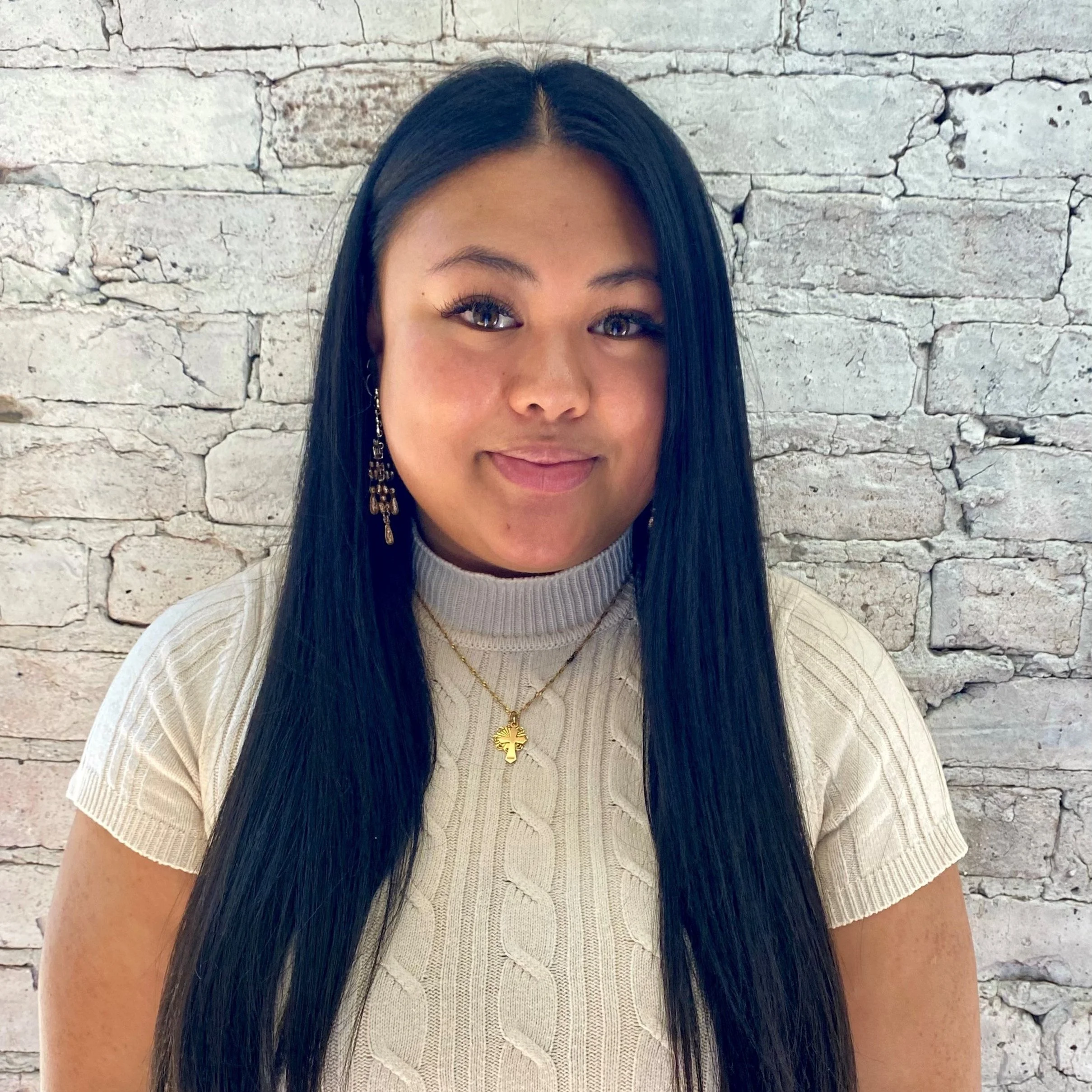 A woman with long black hair and tan skin wearing a beige turtleneck sweater, gold cross necklace, and gold earrings, smiling in front of a white brick wall.