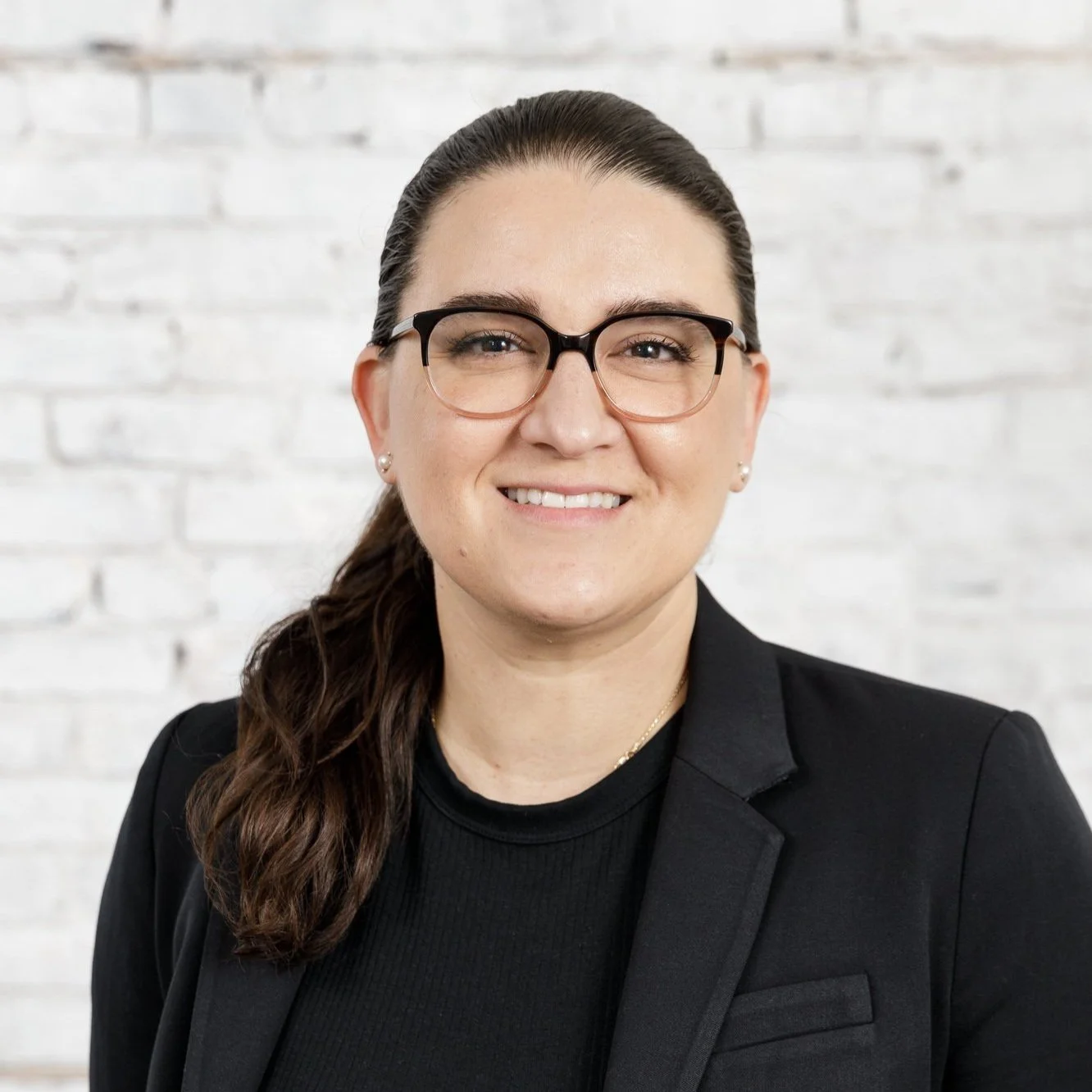 A woman with long dark hair, glasses, and small earrings smiling in front of a white brick wall, wearing a black blazer and black shirt.