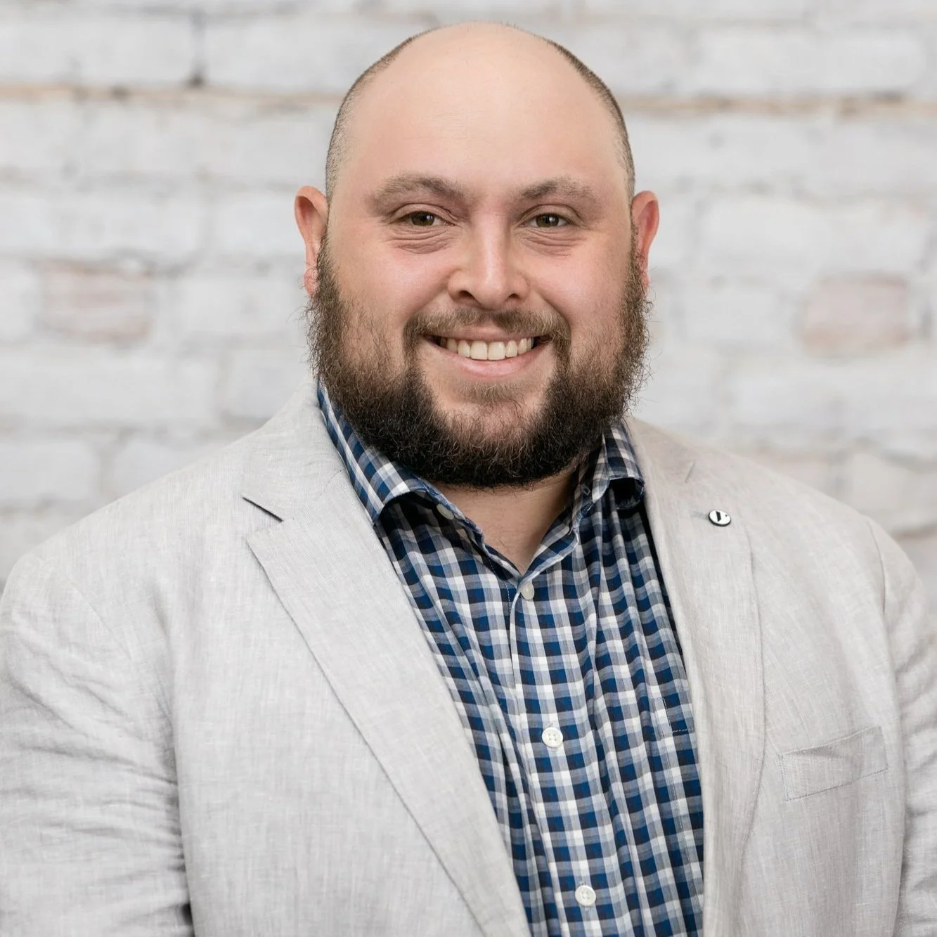 Man with a beard and short hair smiling, wearing a light gray blazer over a blue checkered shirt, against a white brick wall background.