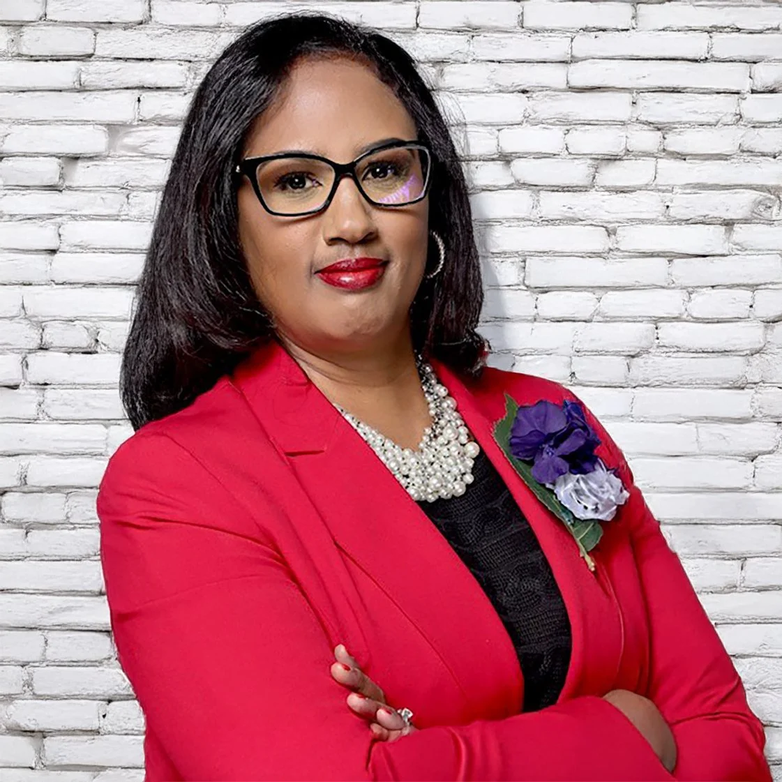 A woman in a red blazer and black top with a purple, white, and green flower brooch, wearing glasses and a pearl necklace, standing with arms crossed against a white brick wall.
