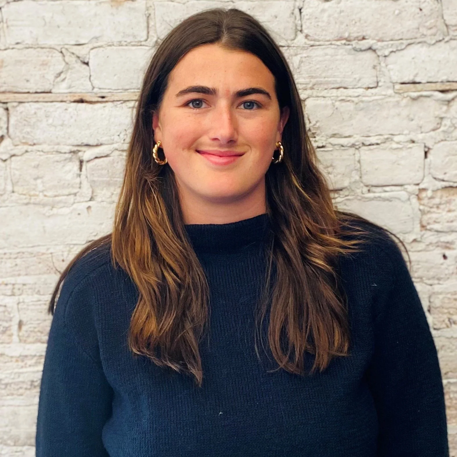 A young woman with shoulder-length brown hair, wearing a black sweater and gold hoop earrings, standing in front of a white brick wall and smiling.