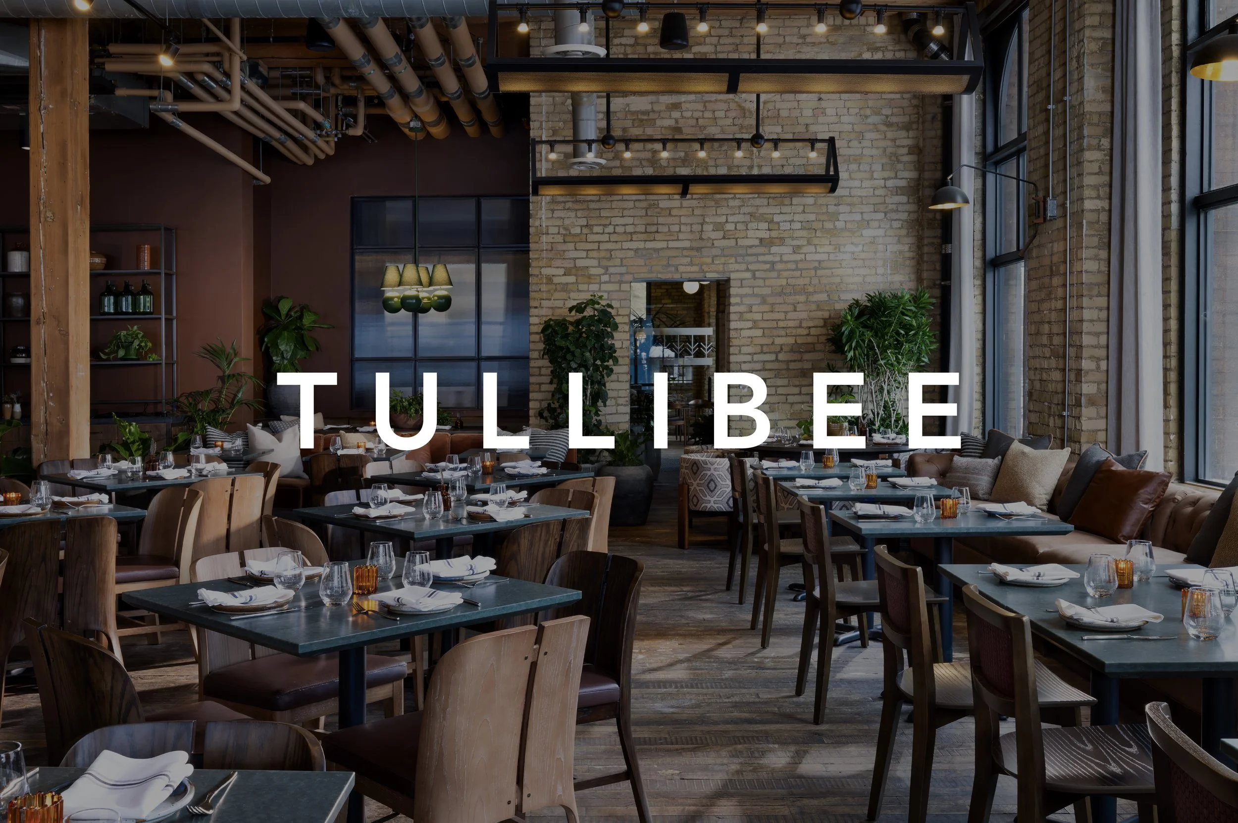 Interior of a restaurant with a wooden dividing wall, glassware hanging upside down, and a leather banquette with tables and chairs, with the word 'TULLIBEE' overlaid.