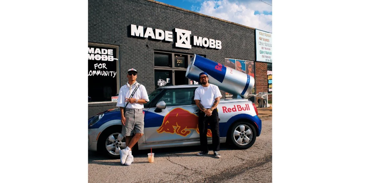 Two men standing in front of a Red Bull car and a clothing store 
