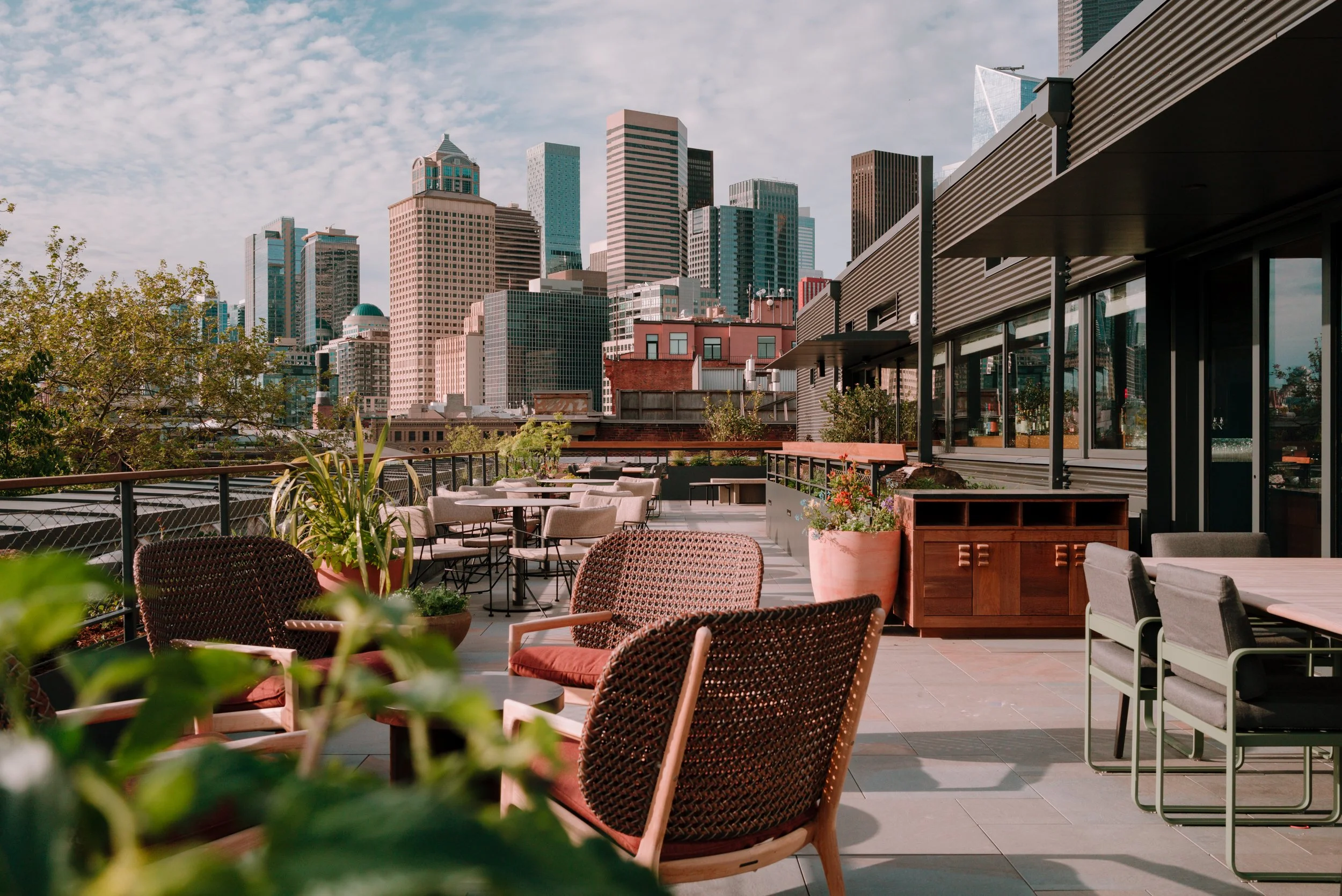 A white stucco building with blue awnings and decorative architectural details, set against tall, modern apartment buildings, with a small front garden, a car parked on the street, and trees providing partial shade.