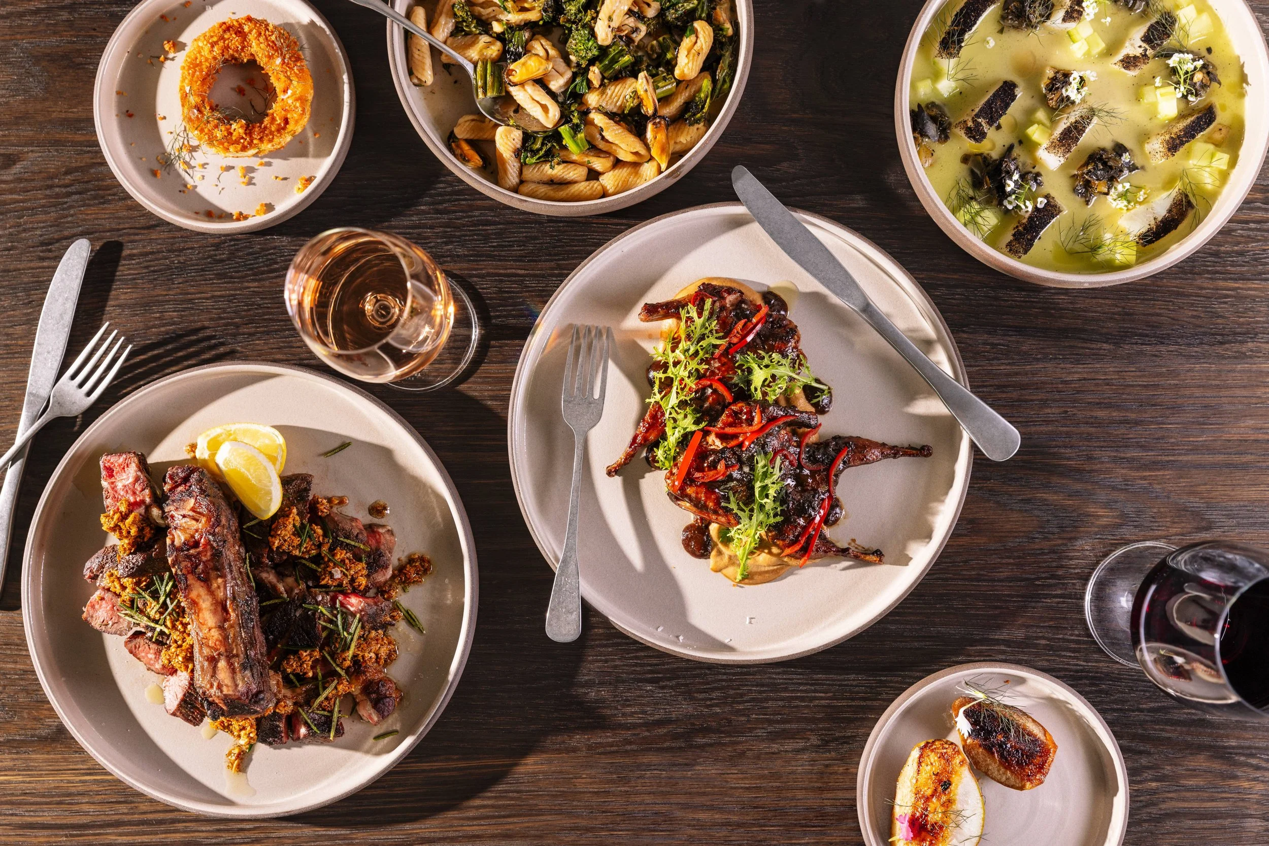 A table with various dishes including a plate of roasted meat with lemon slices, a plate with grilled chicken topped with herbs, a bowl of pasta with greens, a bowl of soup with vegetables and seaweed, and a small dish with grilled bread, along with a glass of rosé and a glass of red wine.