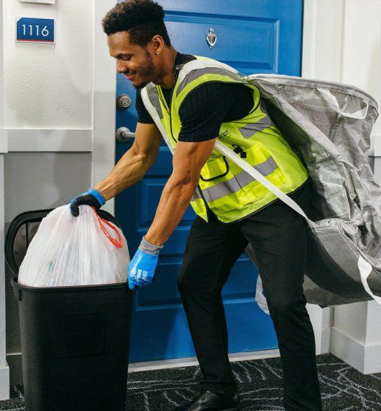 A man wearing a high visibility vest and gloves is throwing a plastic bag into a black trash can near a blue door with the number 1116.
