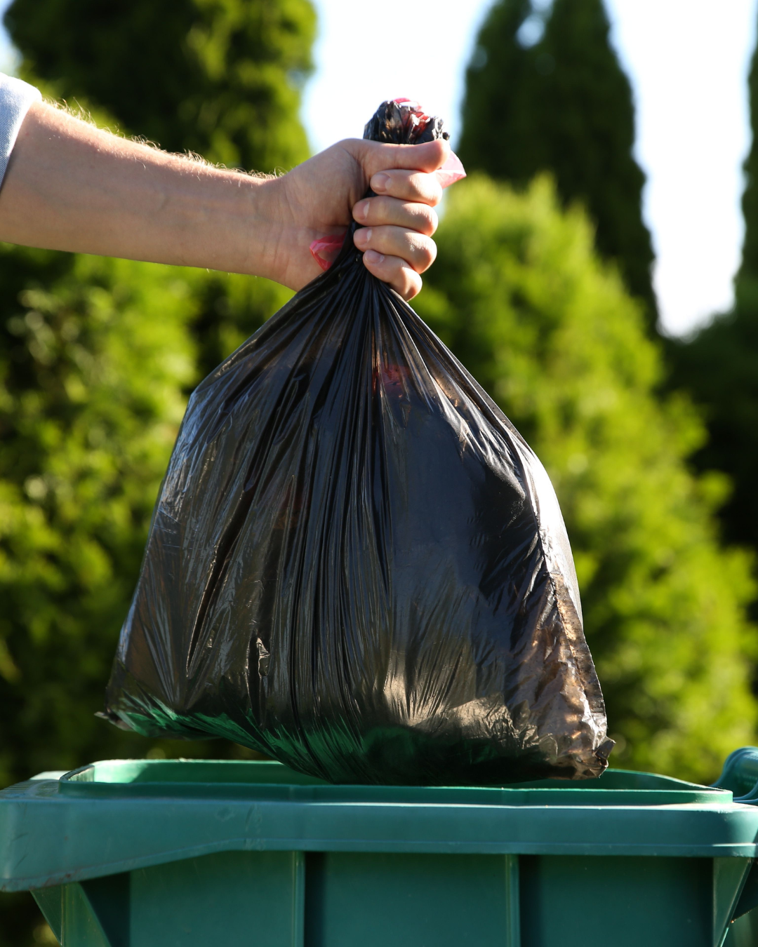 A person's hand throwing a black trash bag into a green trash bin outdoors with green trees and blue sky in the background.