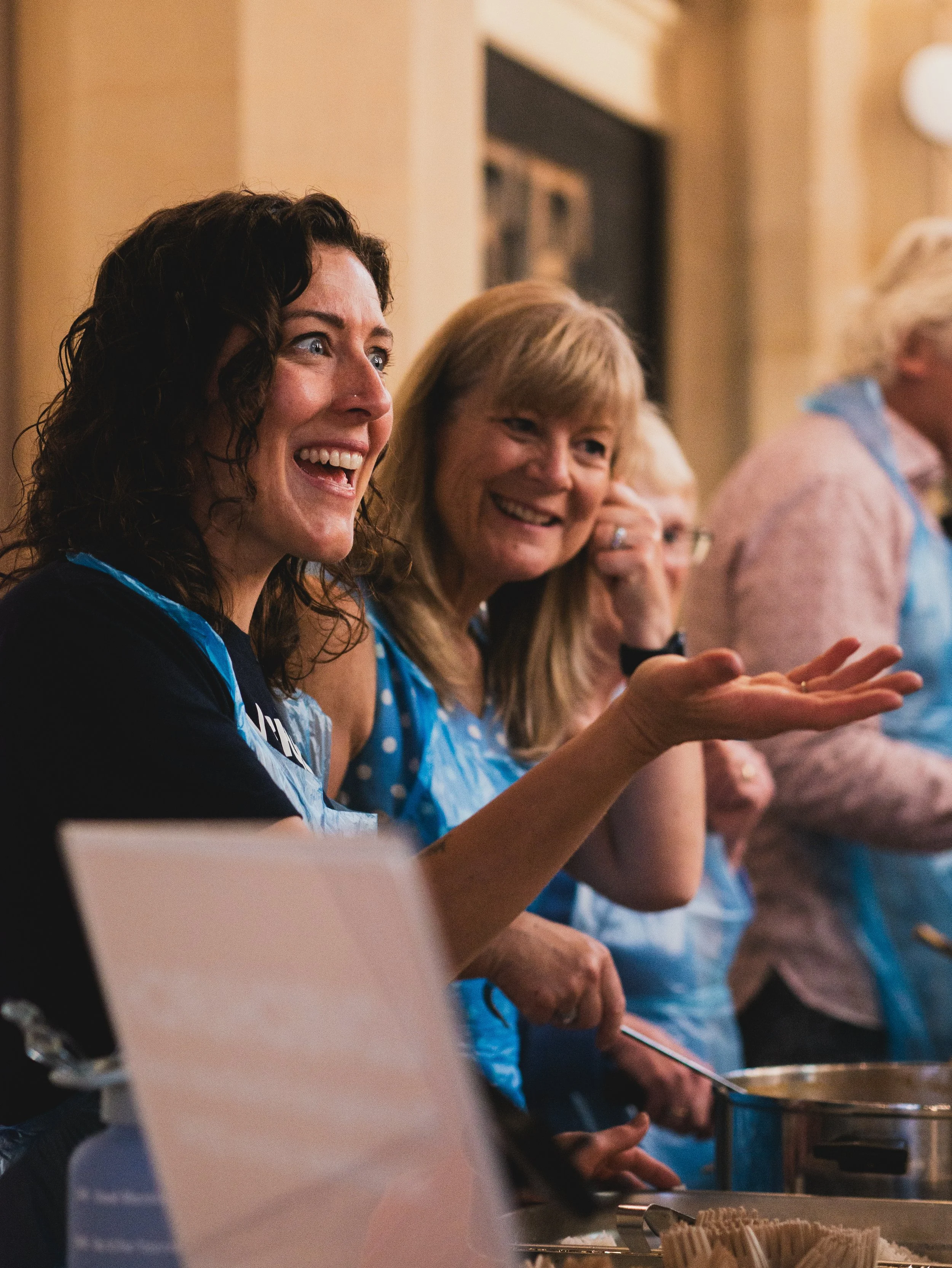 Women smiling and cooking together in a kitchen, wearing blue aprons.