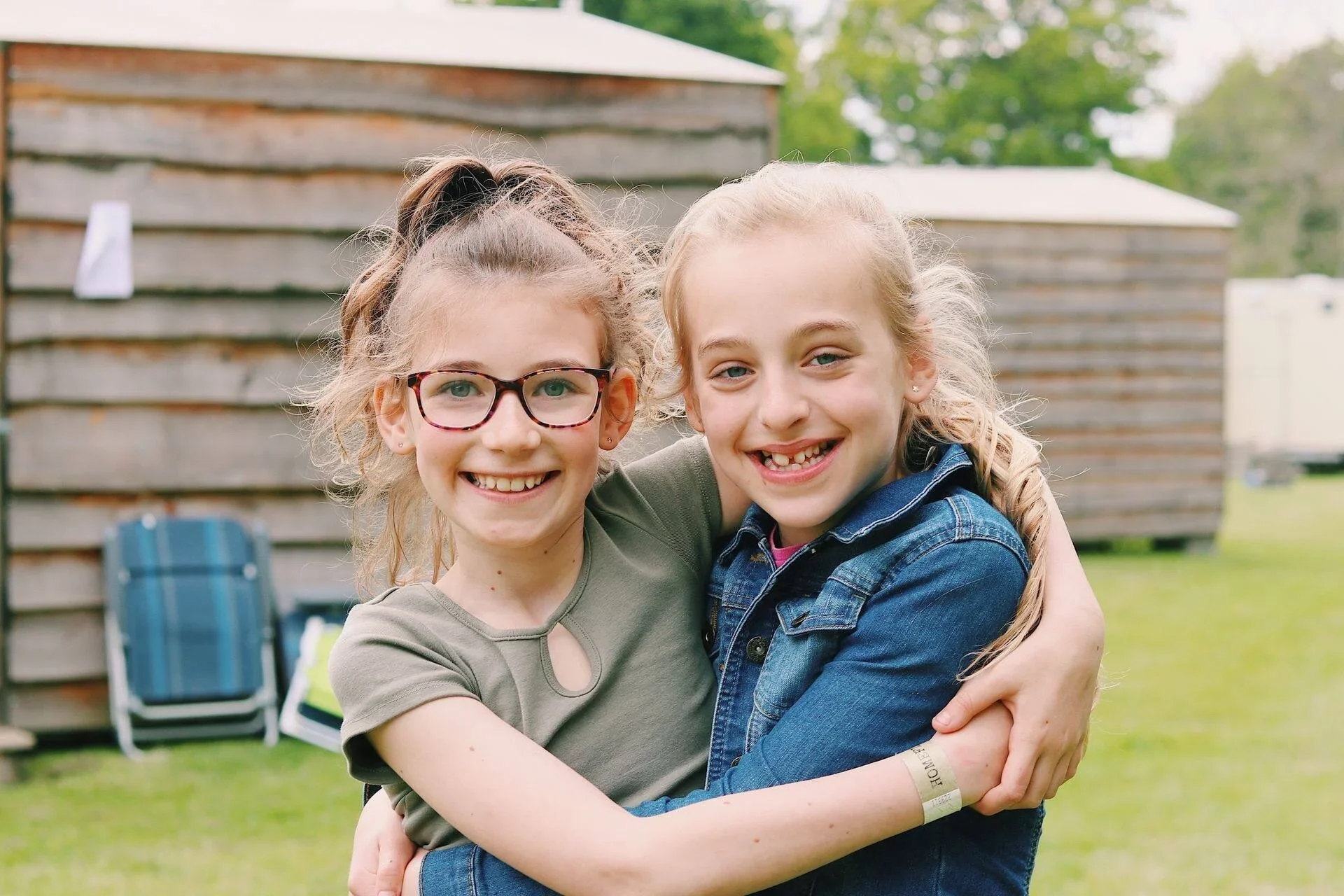 Two young girls with braces hugging and smiling outdoors, with a wooden shed and green trees in the background.