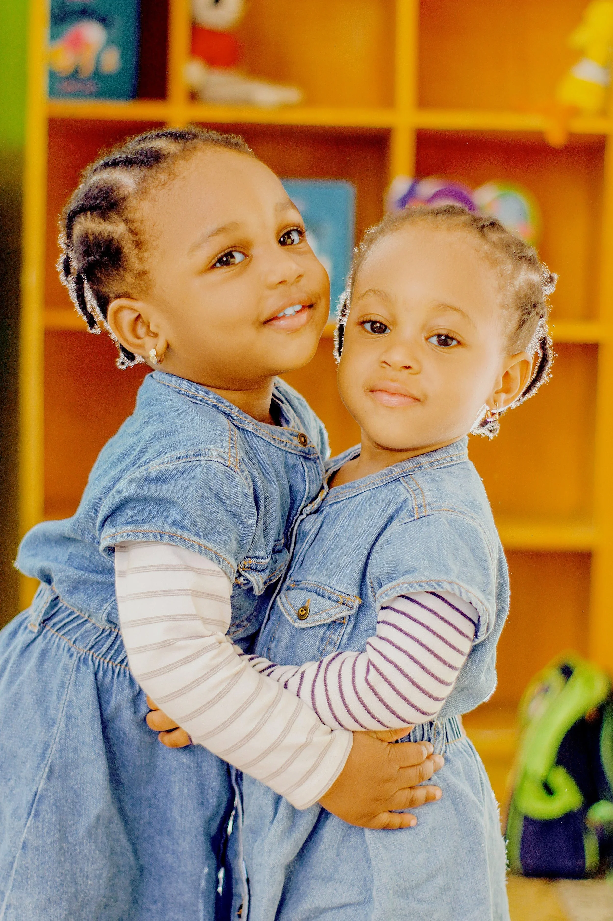 Two young girls with braided hair and earrings, wearing jean outfits, hugging each other in a colorful room with shelves and toys in the background.
