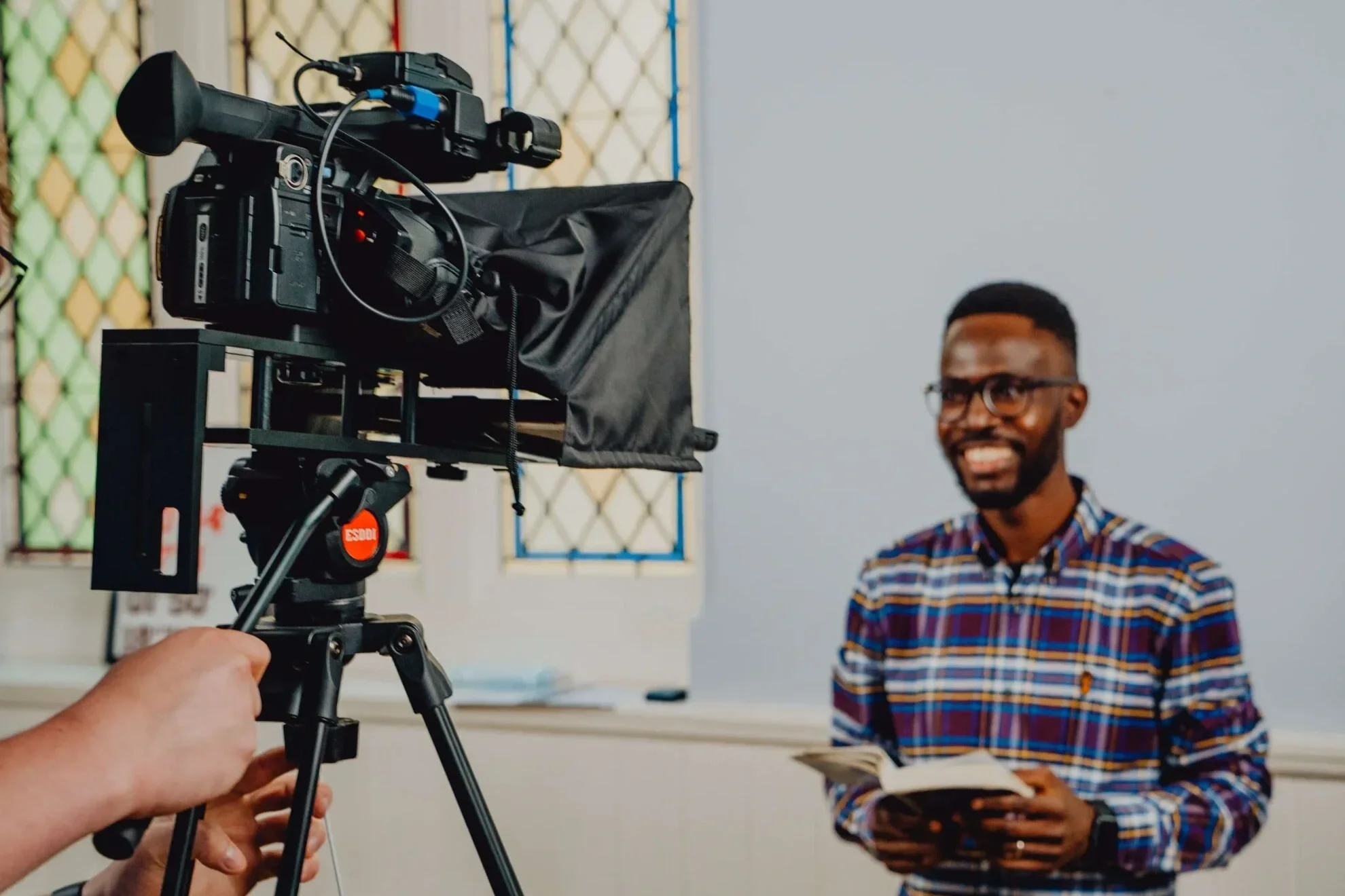 A man with glasses and a plaid shirt is smiling and holding a book while being recorded by a camera on a tripod in a room with a white wall and a window with green, yellow, and red decorative grilles.