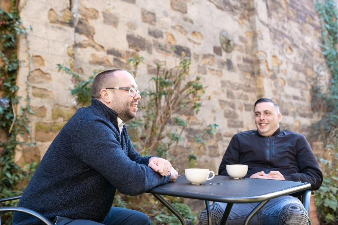 Two men are sitting at a small outdoor table, smiling and chatting, with cups of coffee. They are in a garden with a brick wall and greenery in the background.