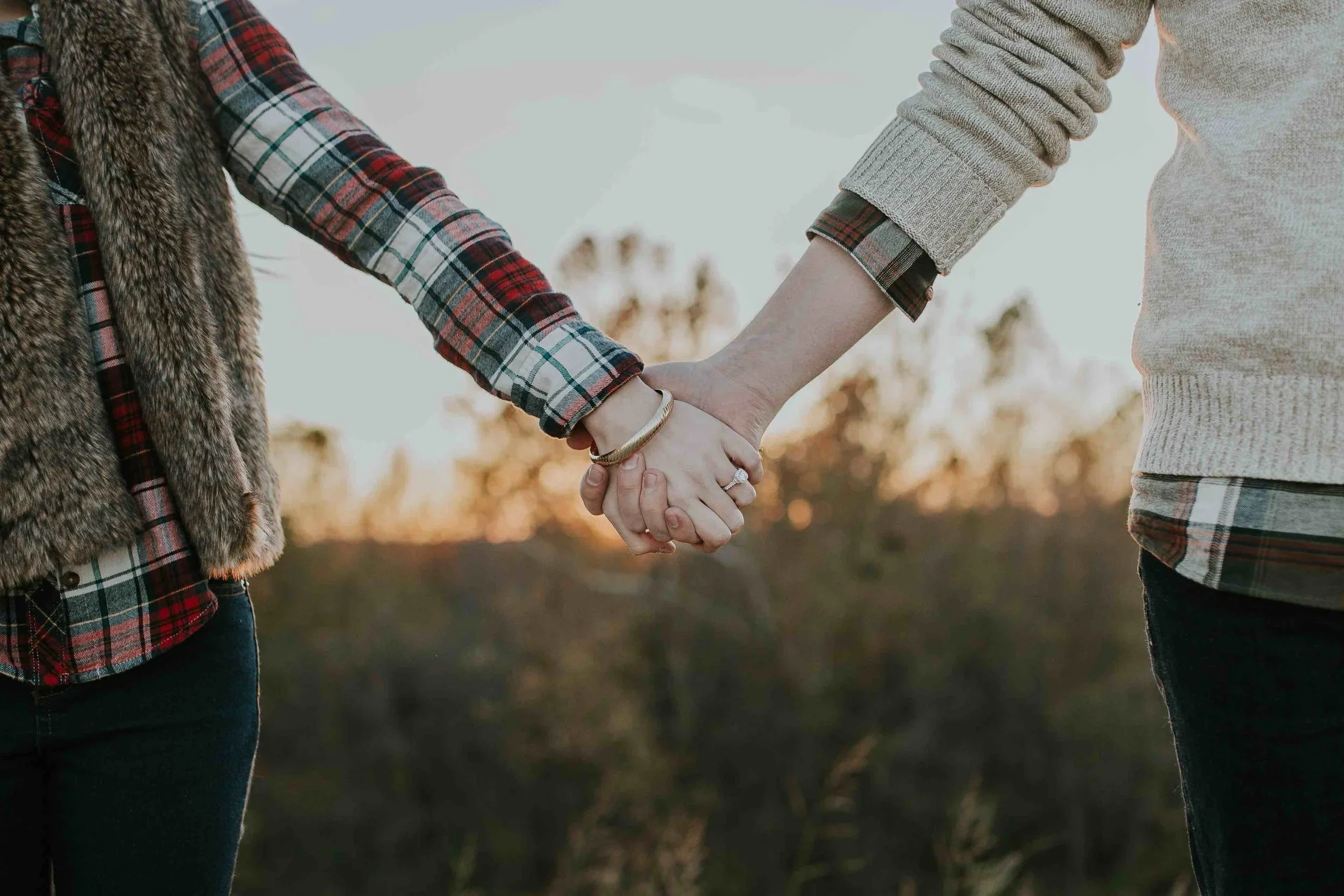 Two people holding hands outdoors during sunset, with trees blurred in the background.