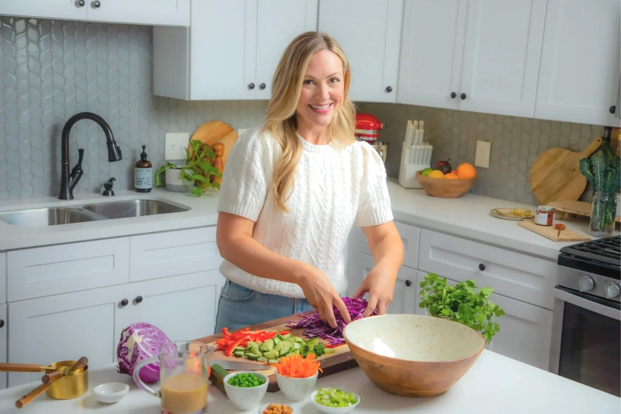 Woman preparing a colorful vegetable salad in a modern kitchen with white cabinets and various vegetables and bowls on the counter.
