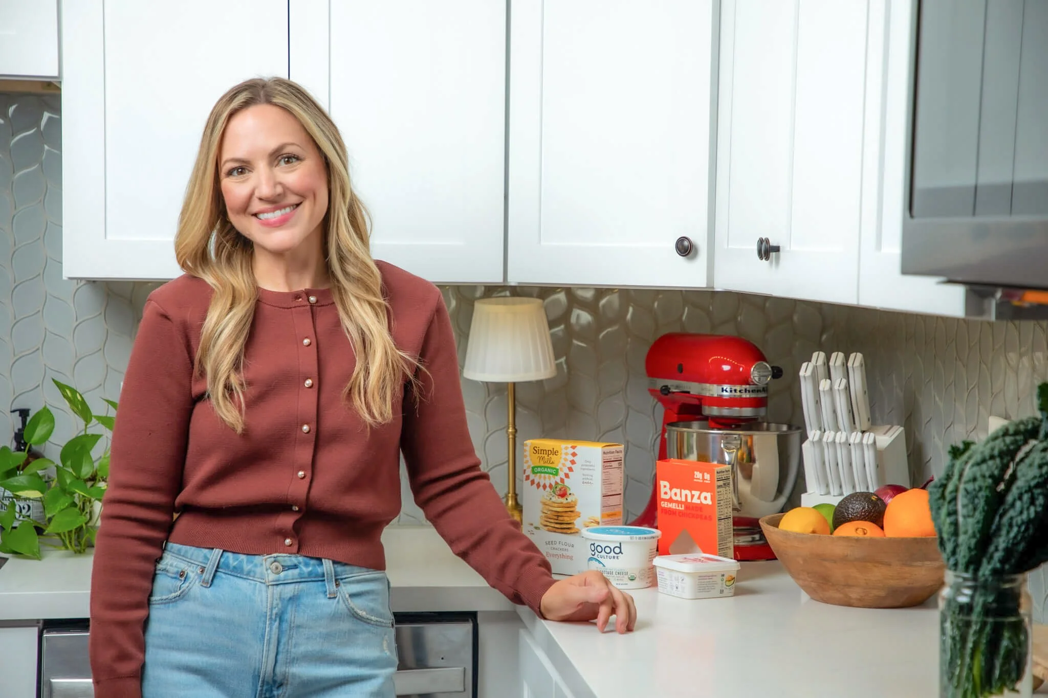 Amy Davis in a kitchen with white cabinets, a red stand mixer, a bowl of fruits, and various food products on the counter.