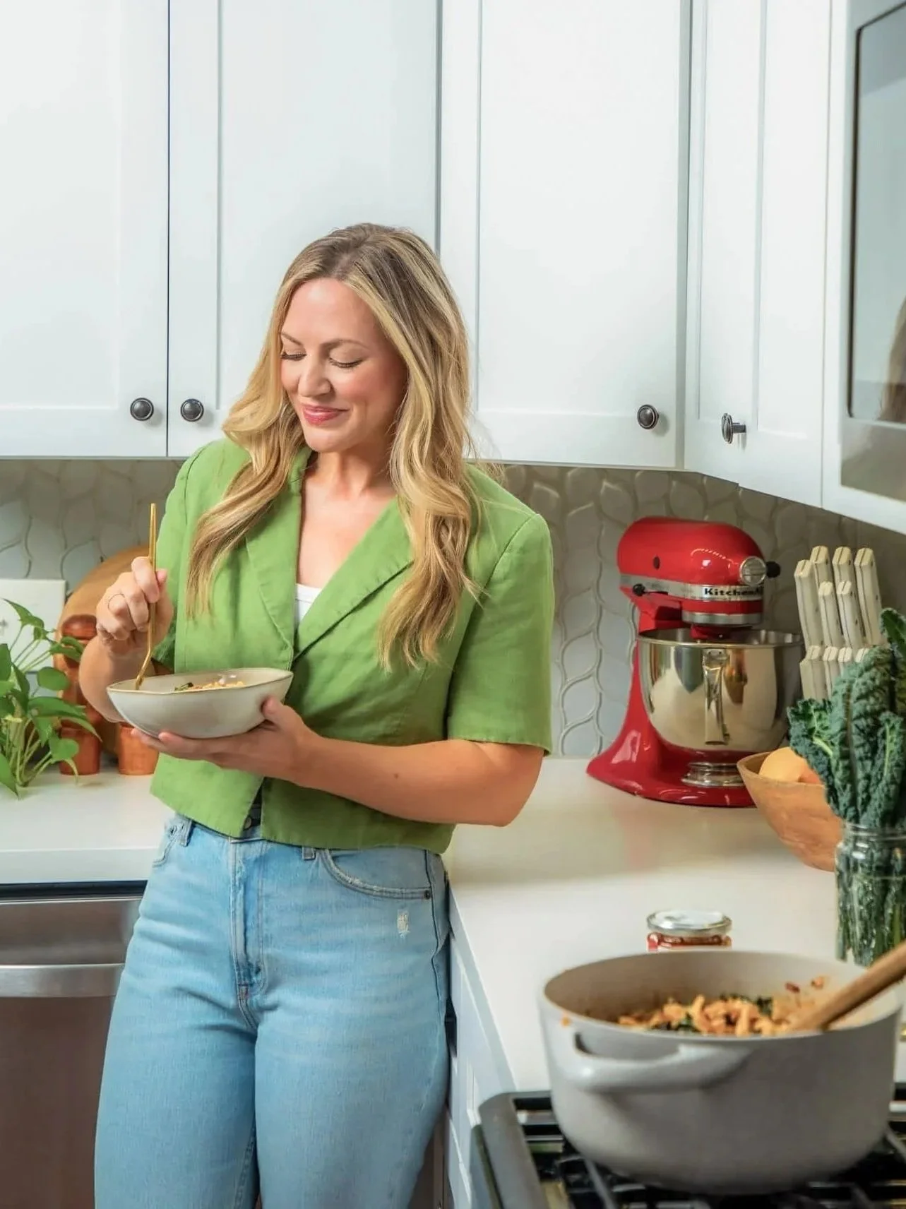 A woman in a green jacket and light blue jeans standing in a kitchen, holding a bowl and eating with a fork, with a red stand mixer and various kitchen items on the counter.