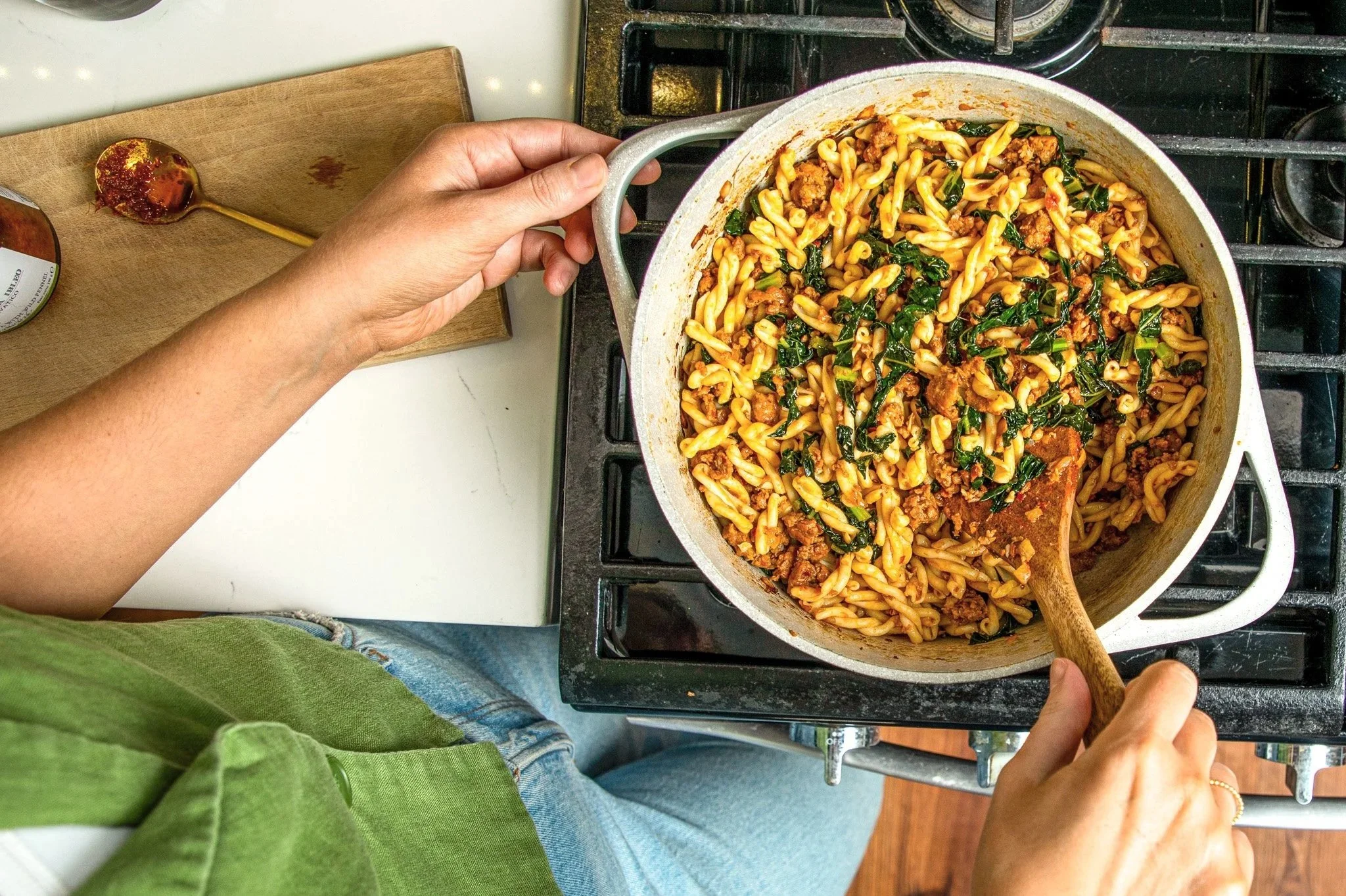 A person cooking a pasta dish with ground meat and leafy greens in a white pot on a stovetop, using a wooden spoon to stir.