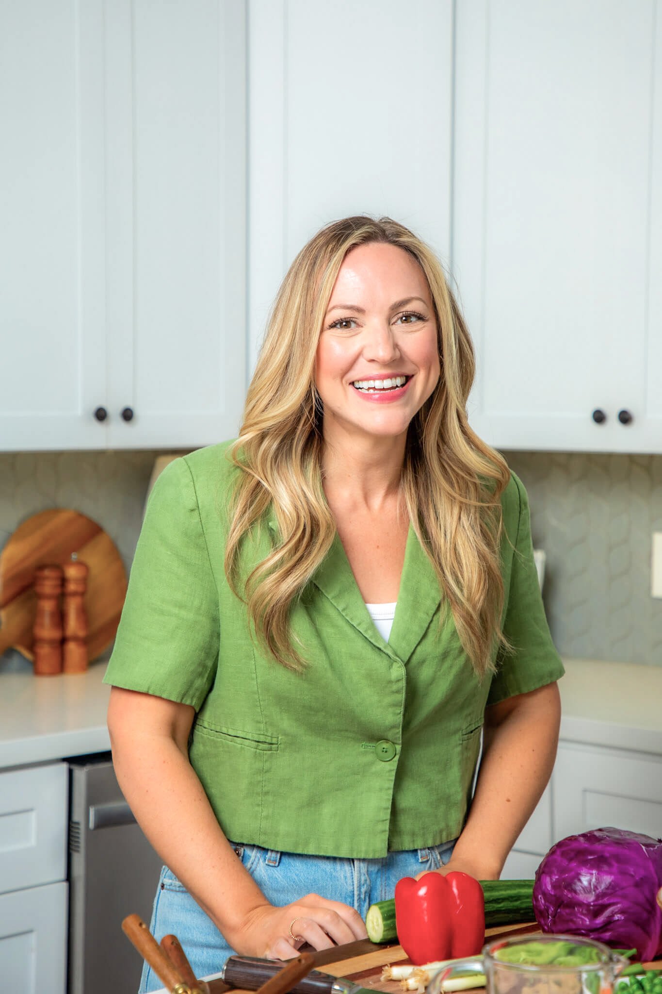 A smiling woman with blonde hair wearing a green shirt and jeans standing in a kitchen with vegetables on the counter.