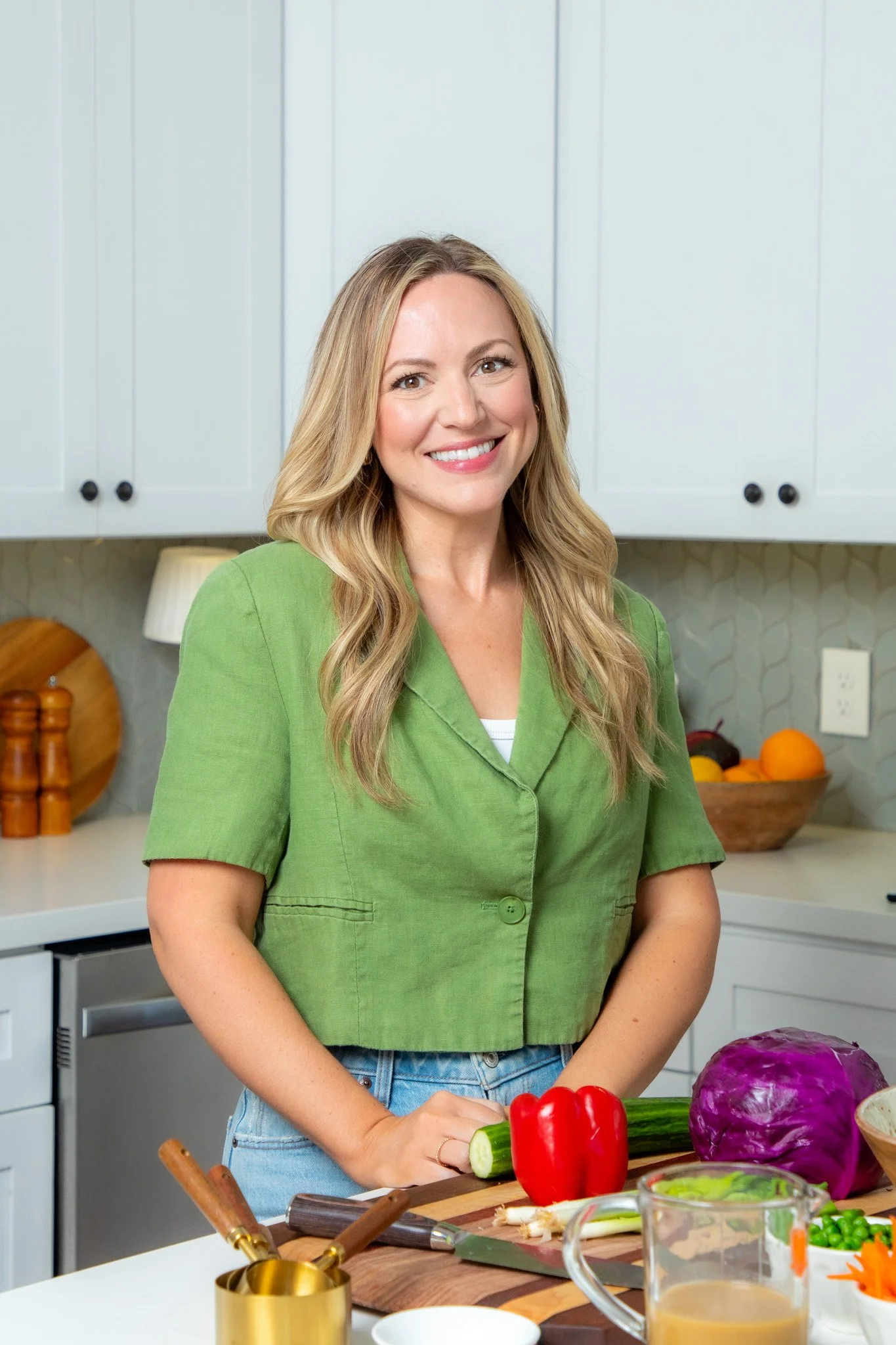 Nutritionist Amy Davis in green shirt smiling in a kitchen with vegetables on the counter, including a red bell pepper, cucumber, purple cabbage, and other produce.