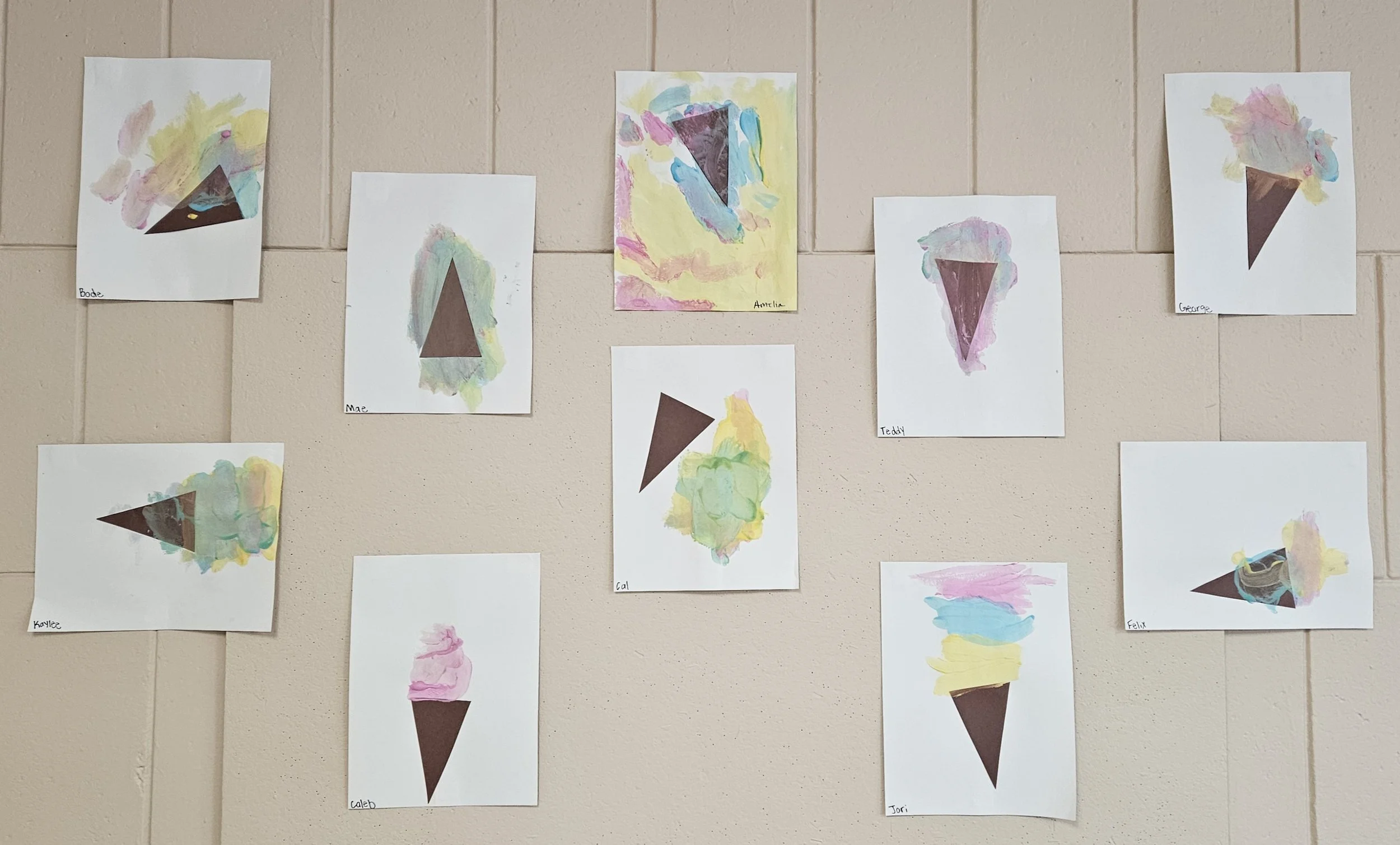 Collection of children's art depicting ice cream cones with colorful, abstract paint on white paper, each labeled with a child's name, displayed on a beige wall.