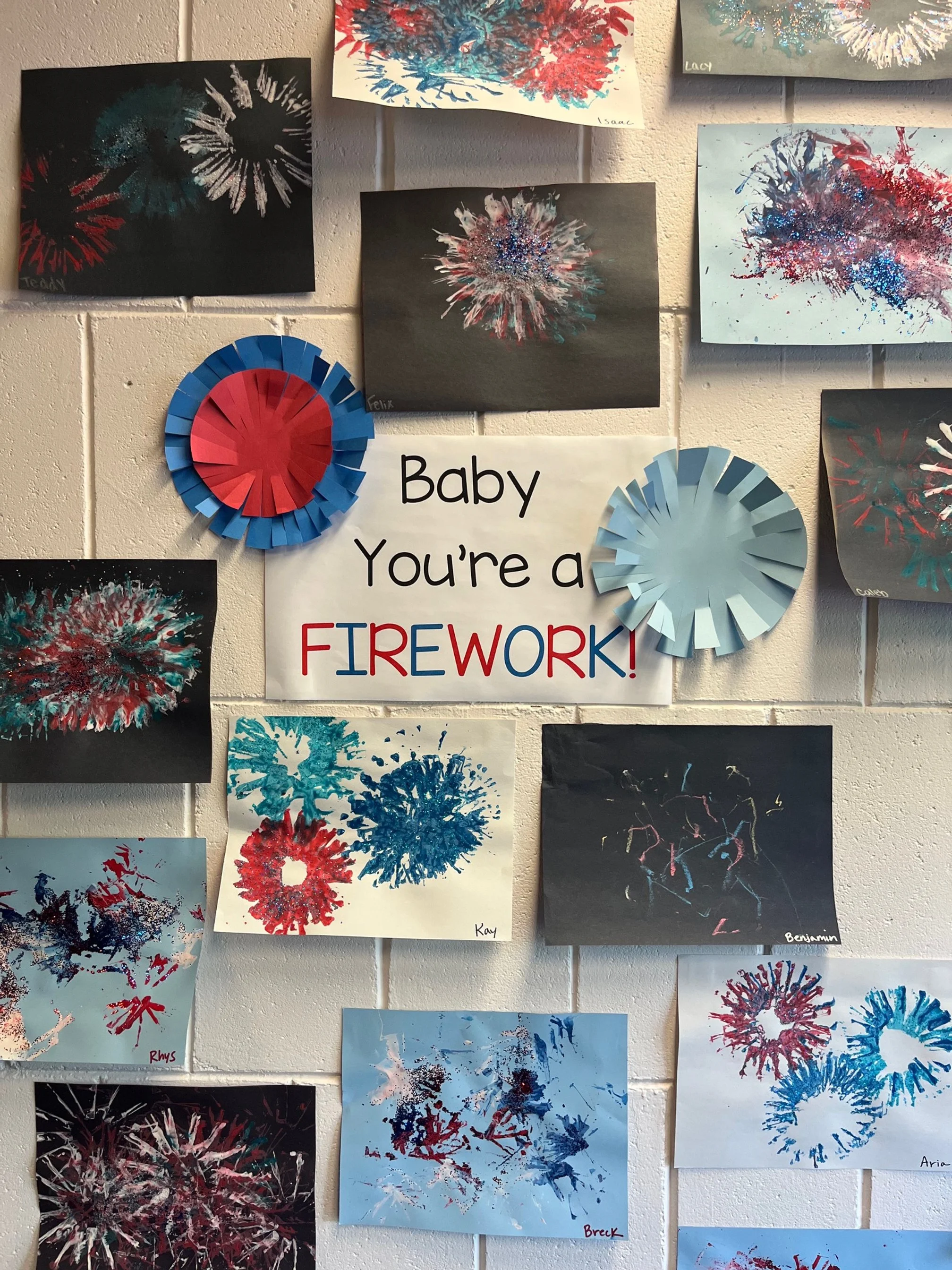 Display of children's artwork depicting colorful firework explosions on paper, with a sign in the center that reads 'Baby You're a FIREWORK!' in black and red letters. Two paper fan decorations resembling firework bursts are also on the wall, one in 