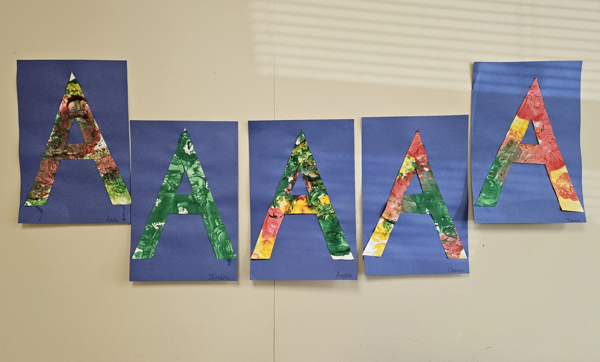 Decorative art display with five blue posters, each with a large, colorful letter A made of marbled paper, and names written at the bottom of each poster.