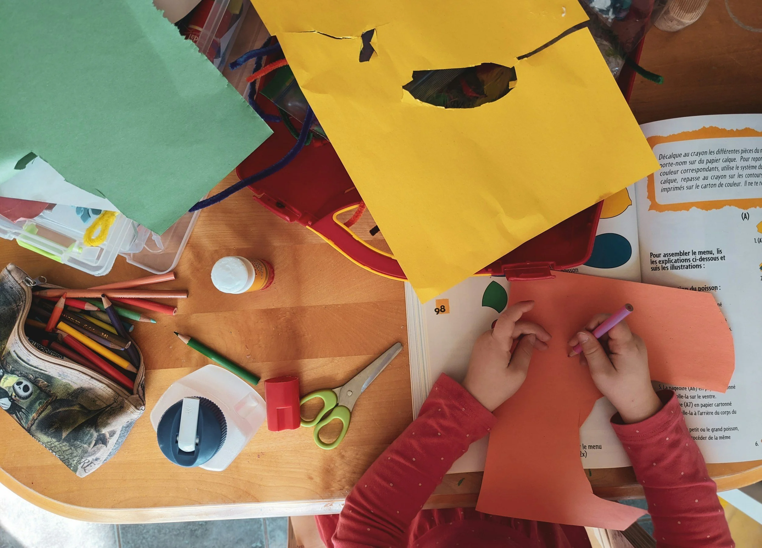 Child creating a craft project with orange construction paper, using markers or crayons, surrounded by school supplies such as scissors, tape, colored paper, and glue on a wooden table.