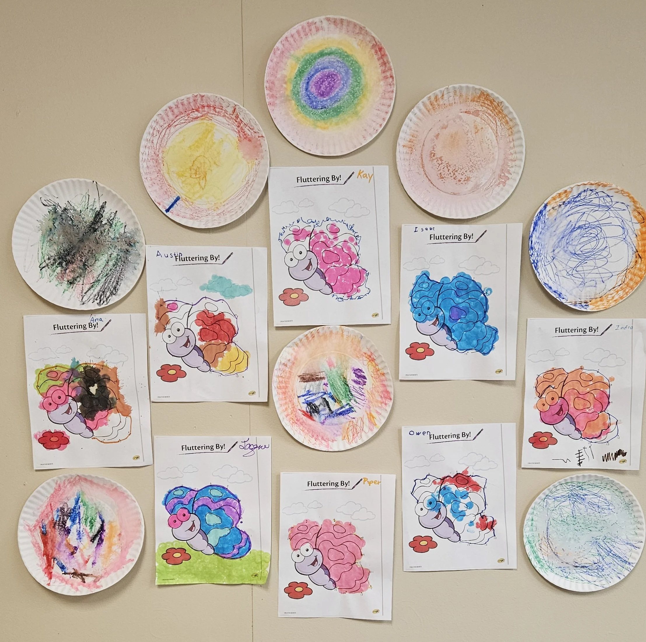 Display of children's artwork on a wall, featuring colorful drawings and painted paper plates with rainbow and abstract designs.