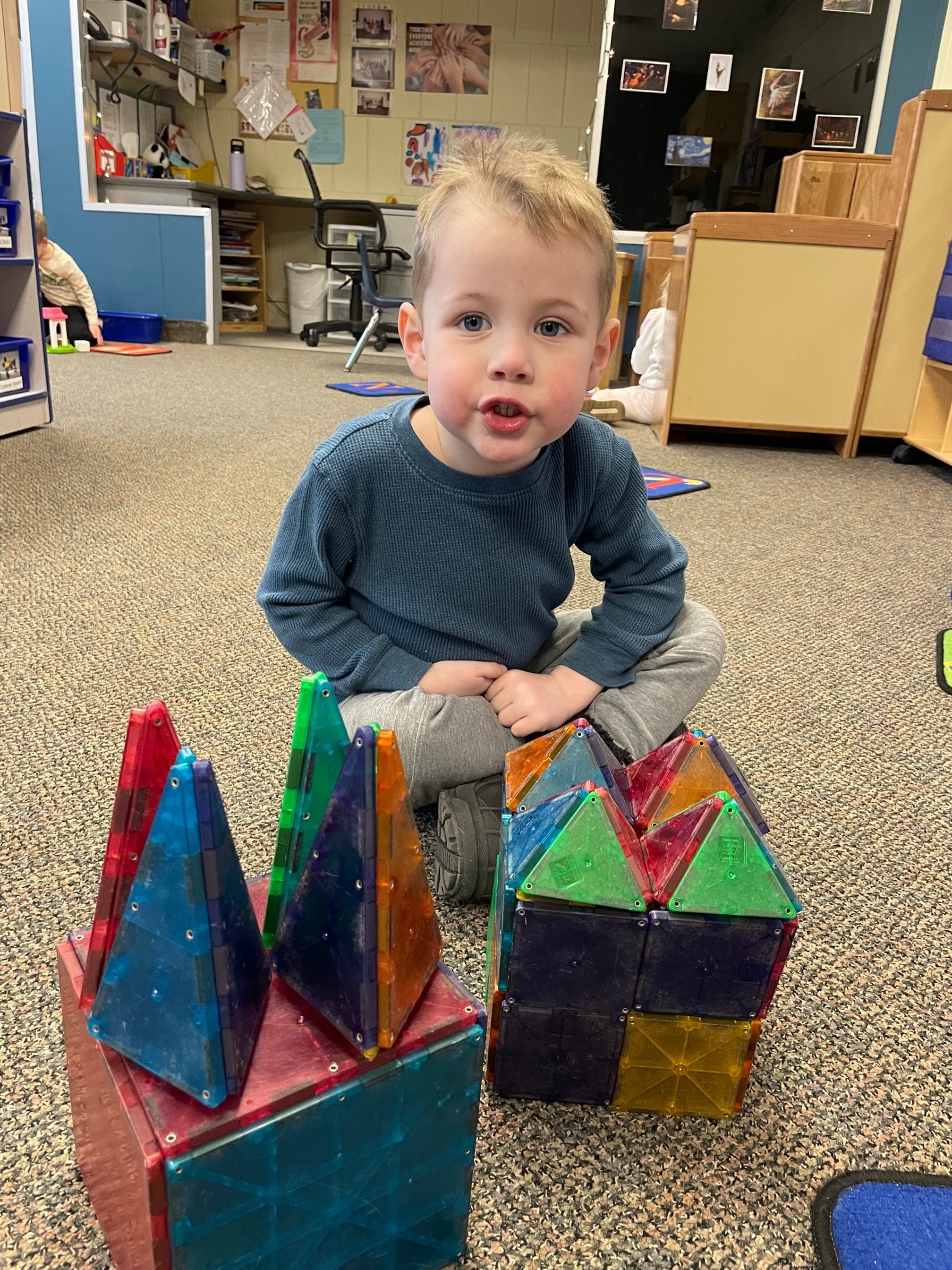 A young boy sitting on the carpeted floor of a classroom, surrounded by colorful magnetic building tiles. The classroom has educational posters, supplies, and other children in the background.