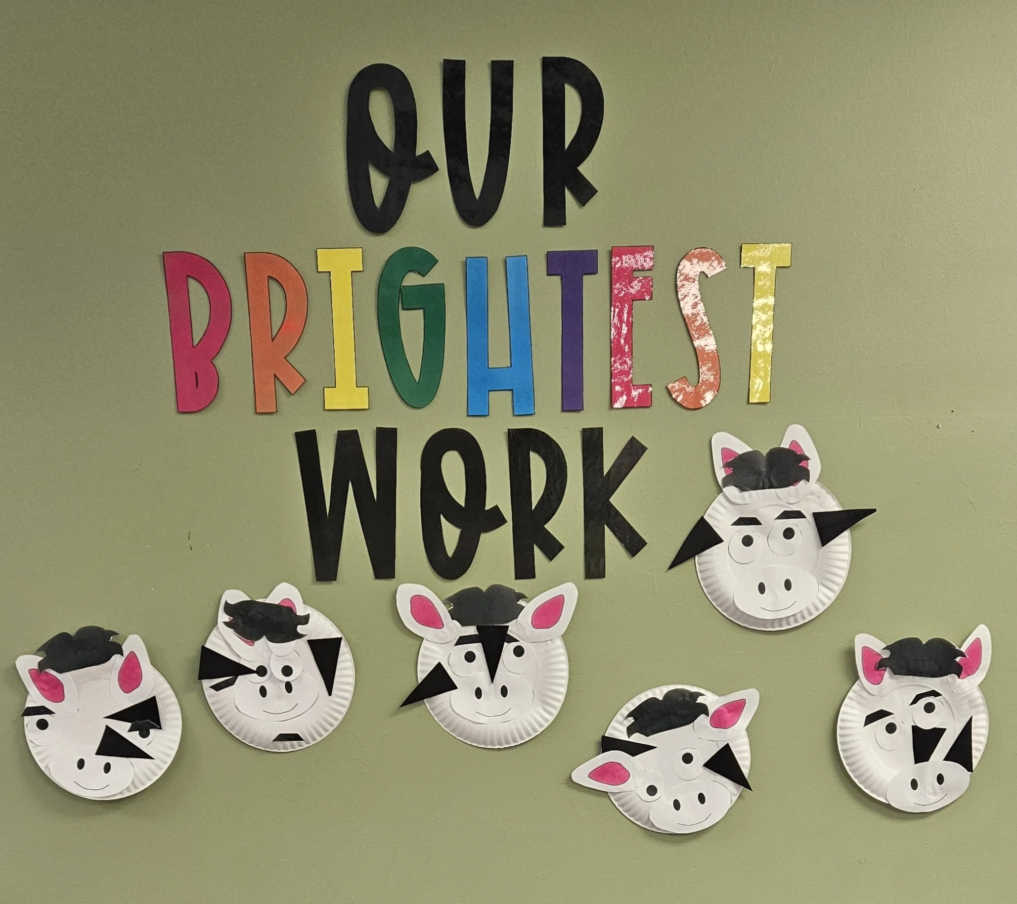 Decorative bulletin board with the phrase 'Our Brightest Work' in colorful letters on a green wall. Below, paper plate animal faces resembling pigs are decorated with black paper hair, ears, eyes, and nostrils.