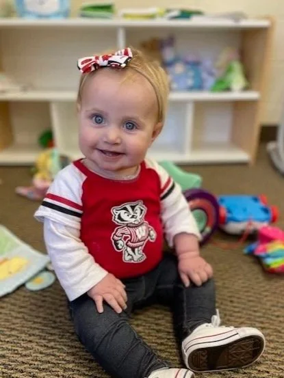 Cute toddler girl with blonde hair, blue eyes, wearing a red and white shirt with a cartoon tiger, dark pants, white shoes, and a red, white, and blue bow in her hair, sitting on a carpeted floor with toys in the background.