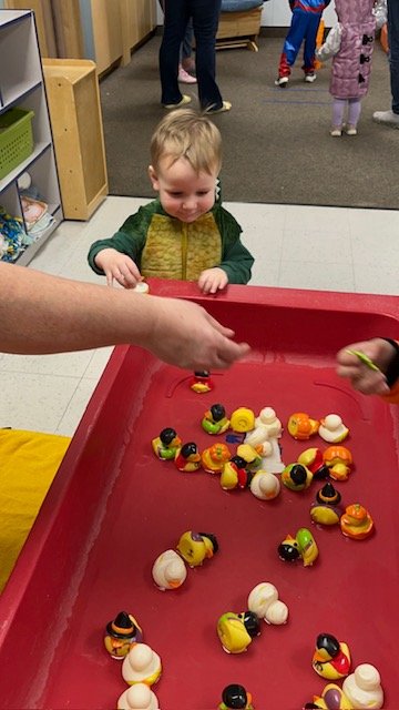 A young boy with blonde hair, wearing a green and yellow jacket, is smiling and watching as someone hands him a plastic duck toy. The boy is standing at a red water table with toy ducks in various colors, including white, yellow, orange, black, and g
