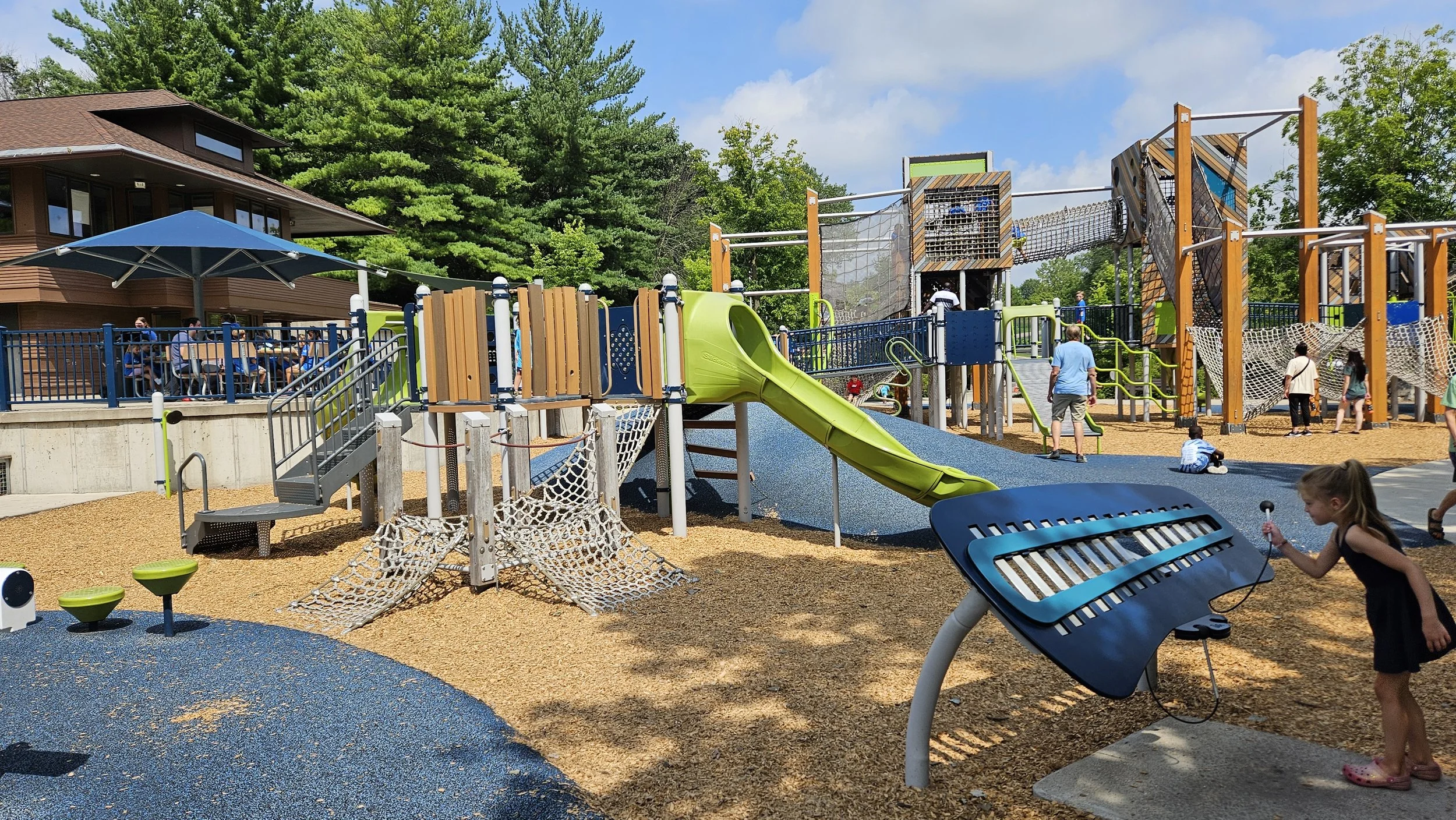 Children playing on a colorful playground with slides, climbing nets, and other play structures, with adults supervising nearby.