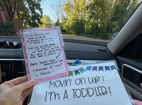 Hand holding two note cards inside a car. One card features handwritten notes with a pink border, and the other has large handwritten text saying, "MOVIN' ON UP! I'M A TODDLER!" with colorful decorations.
