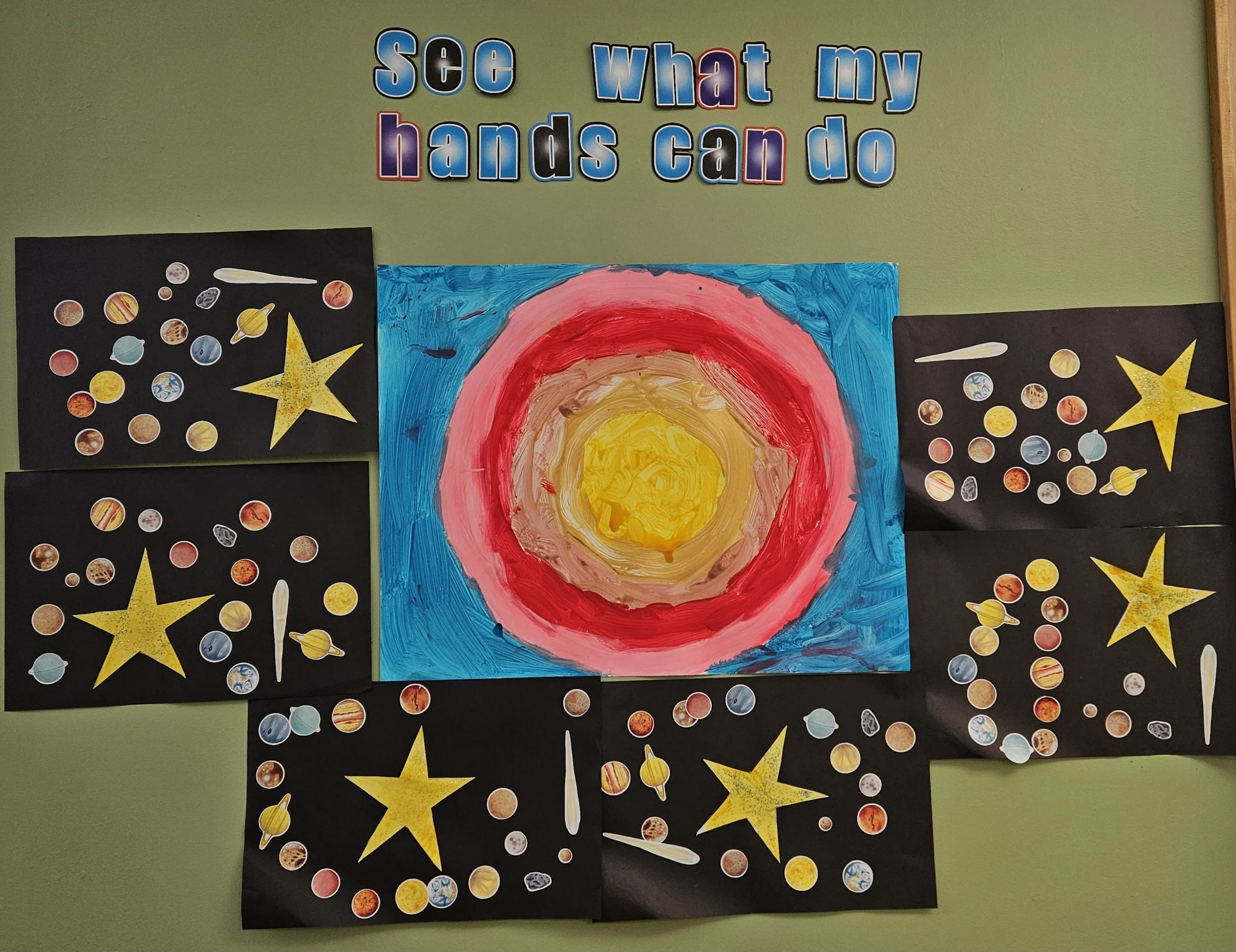 Educational display about what hands can do, featuring a colorful painting of a sun or star in the center, surrounded by black paper with yellow stars, small images of planets and space objects, and the phrase 'see what my hands can do' in multicolor
