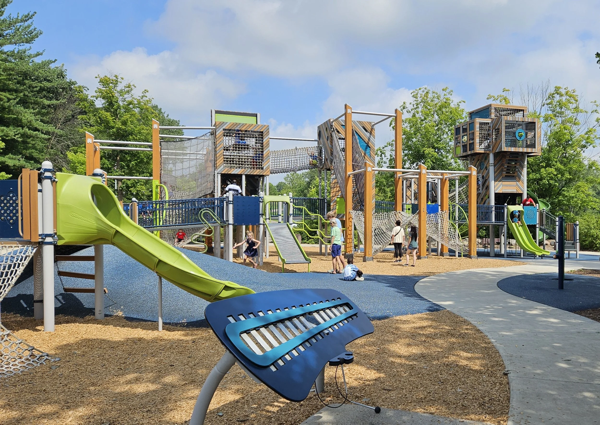 Children playing at a colorful outdoor playground with slides, climbing structures, and balance areas under a partly cloudy sky, surrounded by trees.