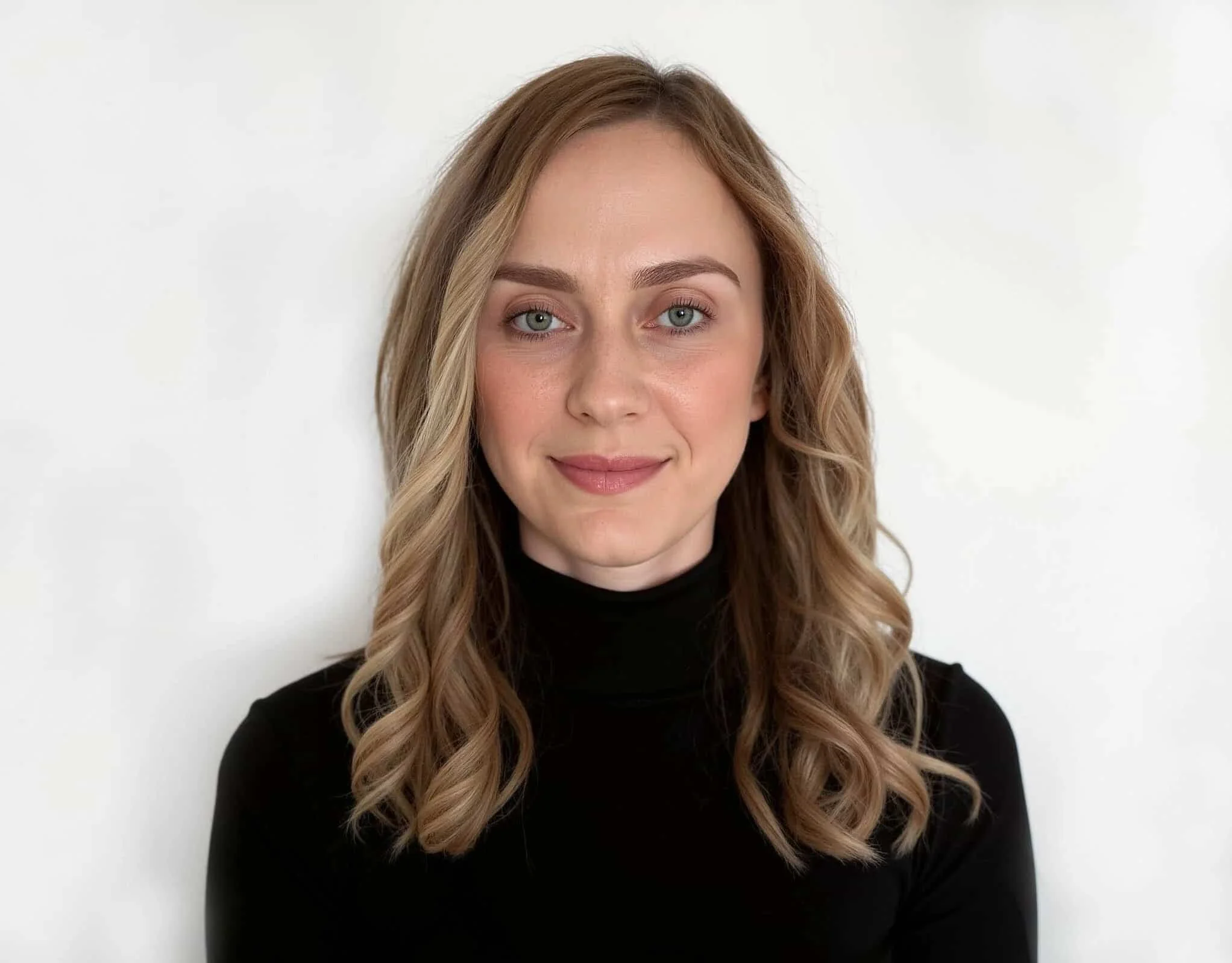 Portrait of a young woman with shoulder-length wavy blonde hair, wearing a black turtleneck, smiling softly against a plain white background.