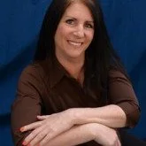 A woman with long dark hair sitting with her arms crossed in front of a blue curtain background.